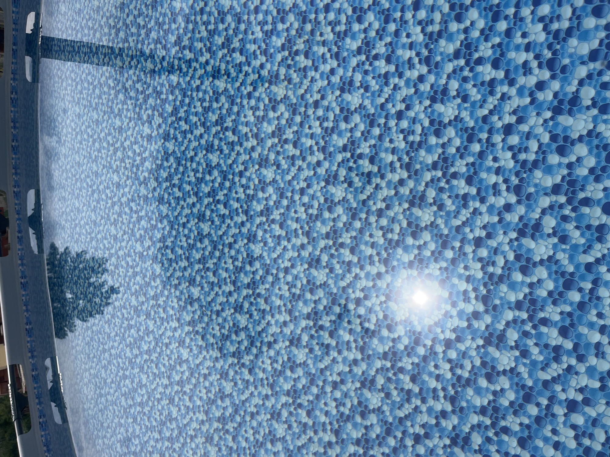 This media file depicts the tiled bottom of an empty swimming pool. The tiles are small and circular, arranged in a mosaic pattern that transitions from lighter blue hues on the left side to darker blues on the right. A prominent sun glare is visible in the upper right portion of the image, indicating a bright, sunny day. On the left side, a shadowy reflection of a tree and what appears to be part of the pool's edge structure are visible. The scene suggests an outdoor setting, likely a residential or public swimming pool, under clear weather conditions. No people or specific activities are discernible. The image is devoid of any visible text. The context provided places this location in Voronezh, Russia.