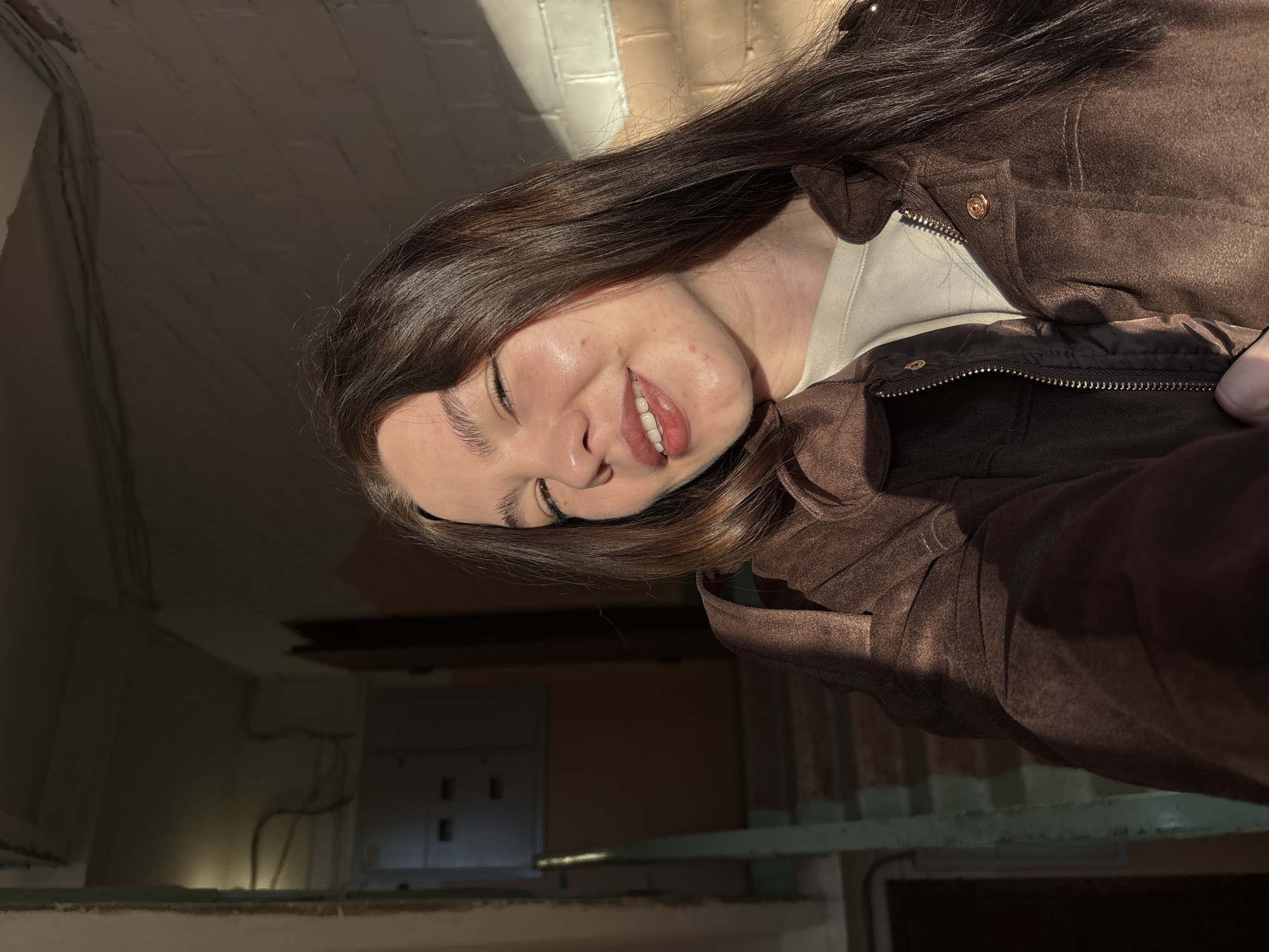 A young woman with long, dark brown hair is captured in a selfie, taken at an angle that rotates the image 90 degrees clockwise. She wears a brown jacket with visible zippers and buttons over a light-colored, possibly cream or white, top. Her head is tilted, and she is smiling broadly with her eyes partially closed, conveying a sense of happiness or contentment. Her light skin is highlighted by direct sunlight.
The setting appears to be an indoor space, possibly a hallway or room within an older building, suggested by the exposed, light-colored brick wall in the background, illuminated by strong sunlight from the upper left. This sunlight casts dramatic shadows, creating a high-contrast scene. The time of day seems to be late afternoon or early morning, indicated by the warm, golden quality of the light. Much of the background is plunged into deep shadow, revealing indistinct shapes, possibly wiring, pipes, or other architectural features. A light-colored horizontal beam or ledge is visible across the lower portion of the image. No visible text is present.