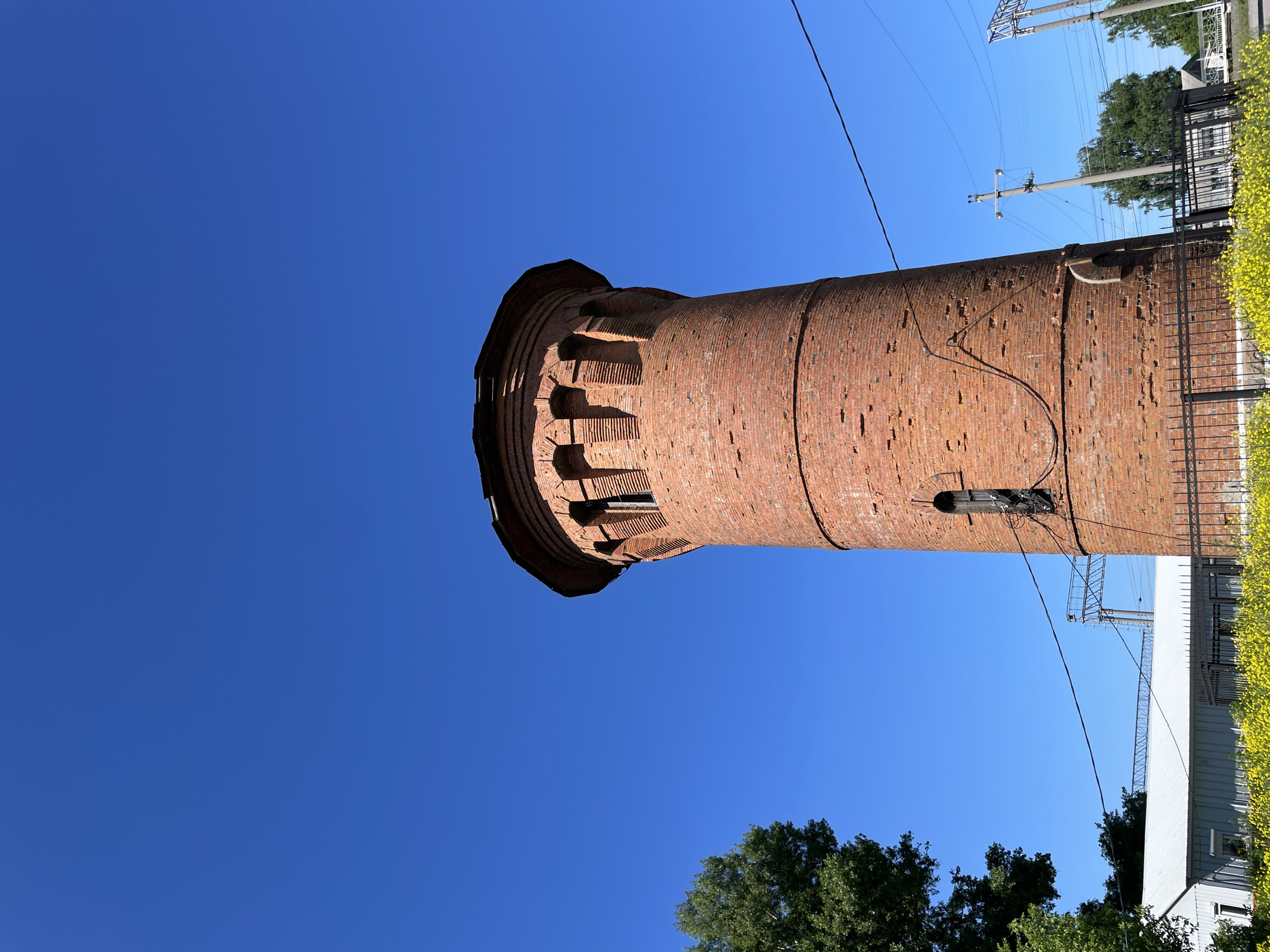 A red brick water tower stands tall against a clear blue sky. The tower has a circular base and tapers slightly towards the top, which is adorned with decorative brickwork and arched openings. A small, dark window is visible on the side of the tower. In the foreground, a black metal fence encloses an area filled with bright yellow flowers. To the left of the tower, a large green tree is partially visible. In the background, there are power lines, a white building with a metal roof, and more trees. The scene appears to be in Artyshta, Russia, as indicated by the information provided.