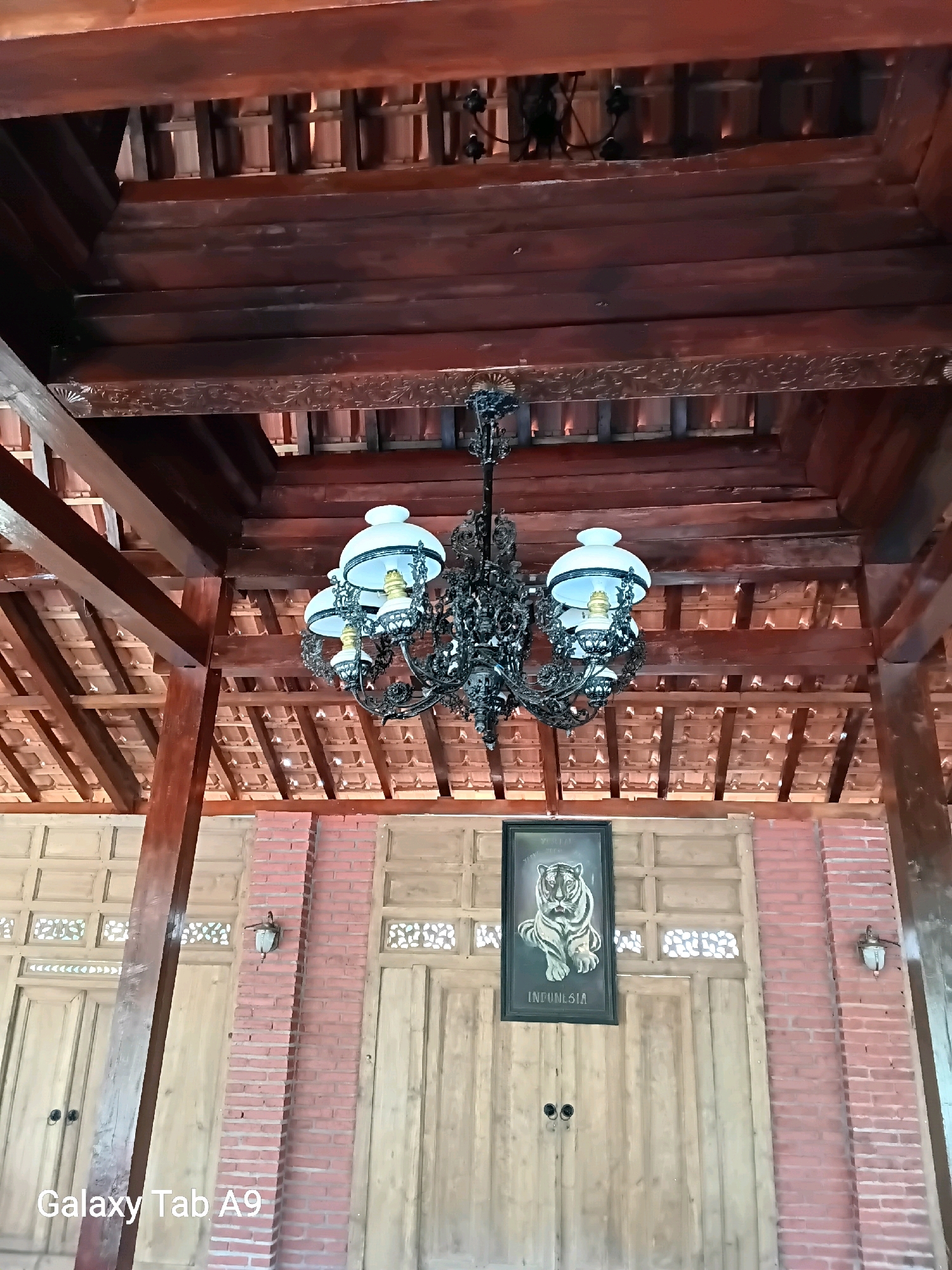 The image shows the interior of a building with a traditional Javanese architectural style. The ceiling is made of dark wood beams and tiles, with a decorative carved wooden frieze. A black metal chandelier with four white glass lamps hangs from the ceiling. Below the ceiling, there are large wooden doors and brick walls. A framed picture of a tiger with the text "INDONESIA" below it is mounted on a wooden door. A single wall-mounted light fixture is visible on a brick column. The overall scene is a snapshot of a room with traditional Indonesian design elements, possibly in Trenggalek, Indonesia. There are no people visible in the image.