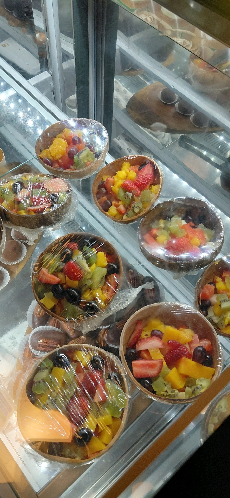 The image shows a display case in a food establishment, likely a bakery or cafe, in Foz do Iguaçu, Brazil. The case is filled with various desserts, primarily fruit salads in small bowls and some chocolate confections. The fruit salads contain chopped strawberries, kiwis, grapes, and what appears to be melon or mango. Several of the fruit salad bowls are wrapped in clear plastic. The display case has glass doors and shelves with internal lighting. In the background, other desserts are visible, including some round, dark-colored pastries and possibly layered cakes. The overall impression is of a clean and well-stocked display of sweet treats.