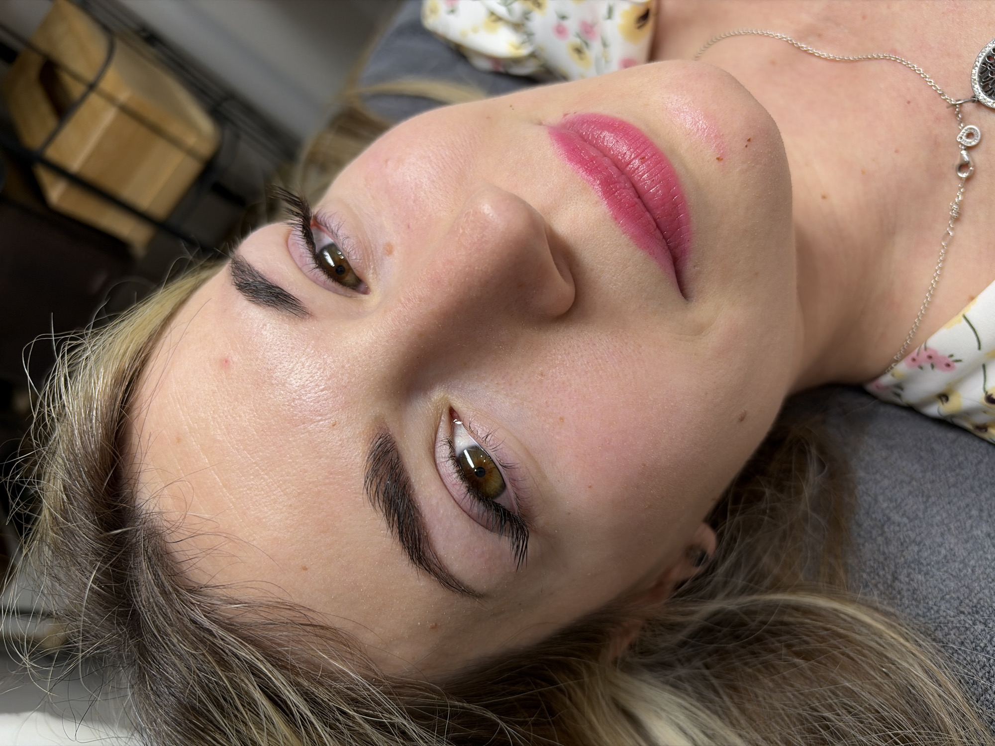 A close-up, tilted shot presents a woman's face. The woman is fair-skinned with hazel eyes, dark eyebrows and eyelashes. Her lips are a bright pink, possibly from lipstick or a cosmetic procedure. She wears a silver chain necklace with a circular pendant. Her hair, a mix of blonde and brown shades, is partially visible in the lower left of the frame. The woman lies on a grey surface, possibly a couch or bed. In the background are floral print fabrics and a wire object. There are no visible landmarks or signs to verify the location being Budva, Montenegro.