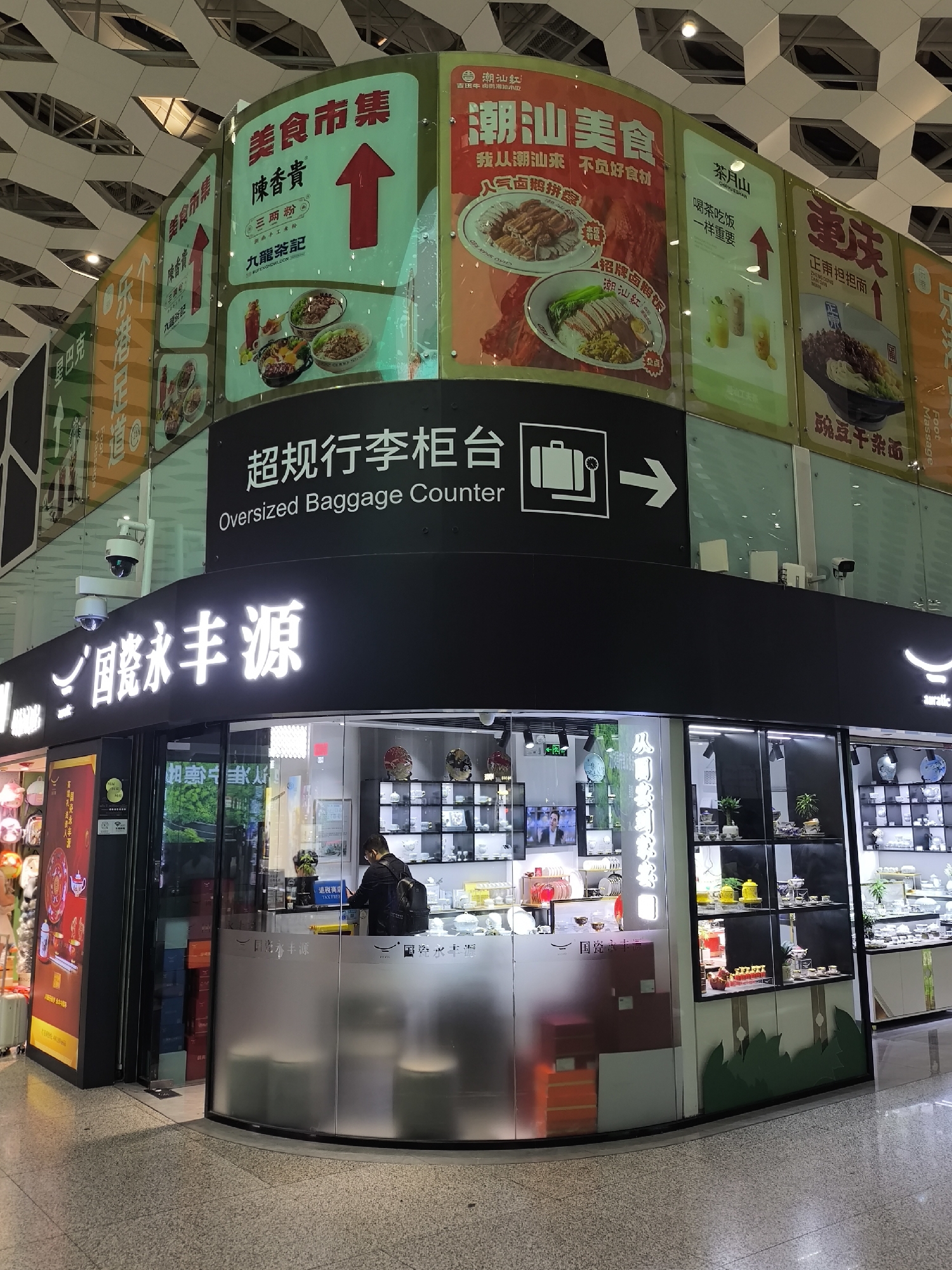 This appears to be an indoor scene, possibly an airport or a large shopping mall in Bao'an District, China. The image shows storefronts and signage in both Chinese and English. There's an "Oversized Baggage Counter" sign with an arrow pointing to the right. There are also advertisements for various food establishments in the background. In the foreground, a storefront displays the characters "国瓷永丰源," which might translate to "National Porcelain Yongfengyuan." A person wearing a backpack is at a counter inside the storefront. The overall environment appears to be clean and modern, with a combination of commercial and infrastructural elements.