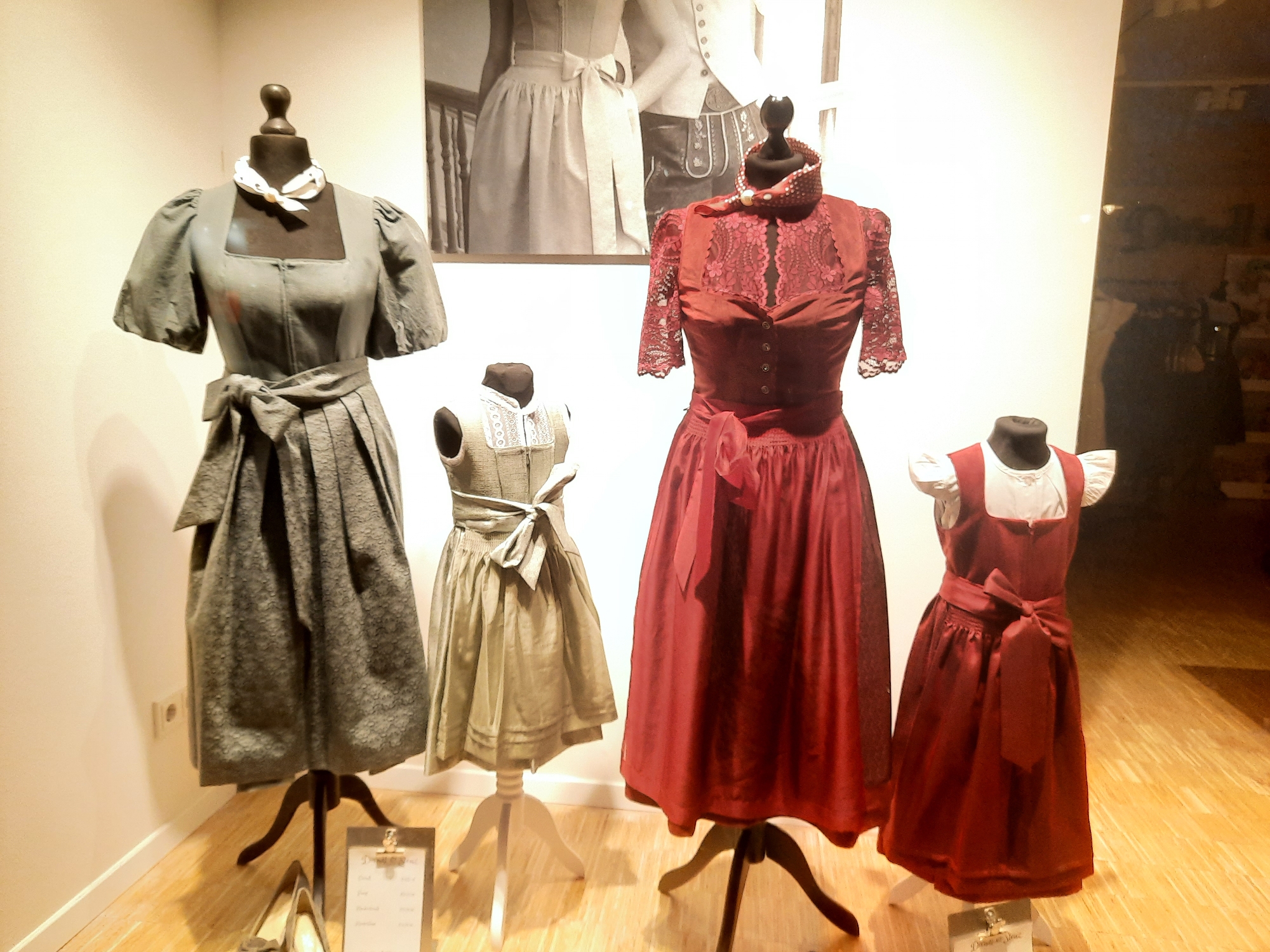 In a retail display, four mannequins are dressed in traditional Bavarian dirndl dresses. The mannequins are arranged in a row on a wooden floor against a plain wall, with a black and white photograph of a couple in traditional attire displayed on the wall behind them. The dirndls vary in color and style. The leftmost dirndl is a dark grey patterned dress with a white scarf. Next to it is a lighter grey, knee-length dirndl with a white apron. The third dirndl from the left is a deep red with intricate lace detailing on the bodice and sleeves, and a matching red ribbon sash. The rightmost dirndl is a dark red pinafore-style dress over a white blouse. A price tag is visible in the foreground, partially obscured. The overall lighting is warm and ambient, typical of a shop window display. This scene is located in the city of Rosenheim, Germany.