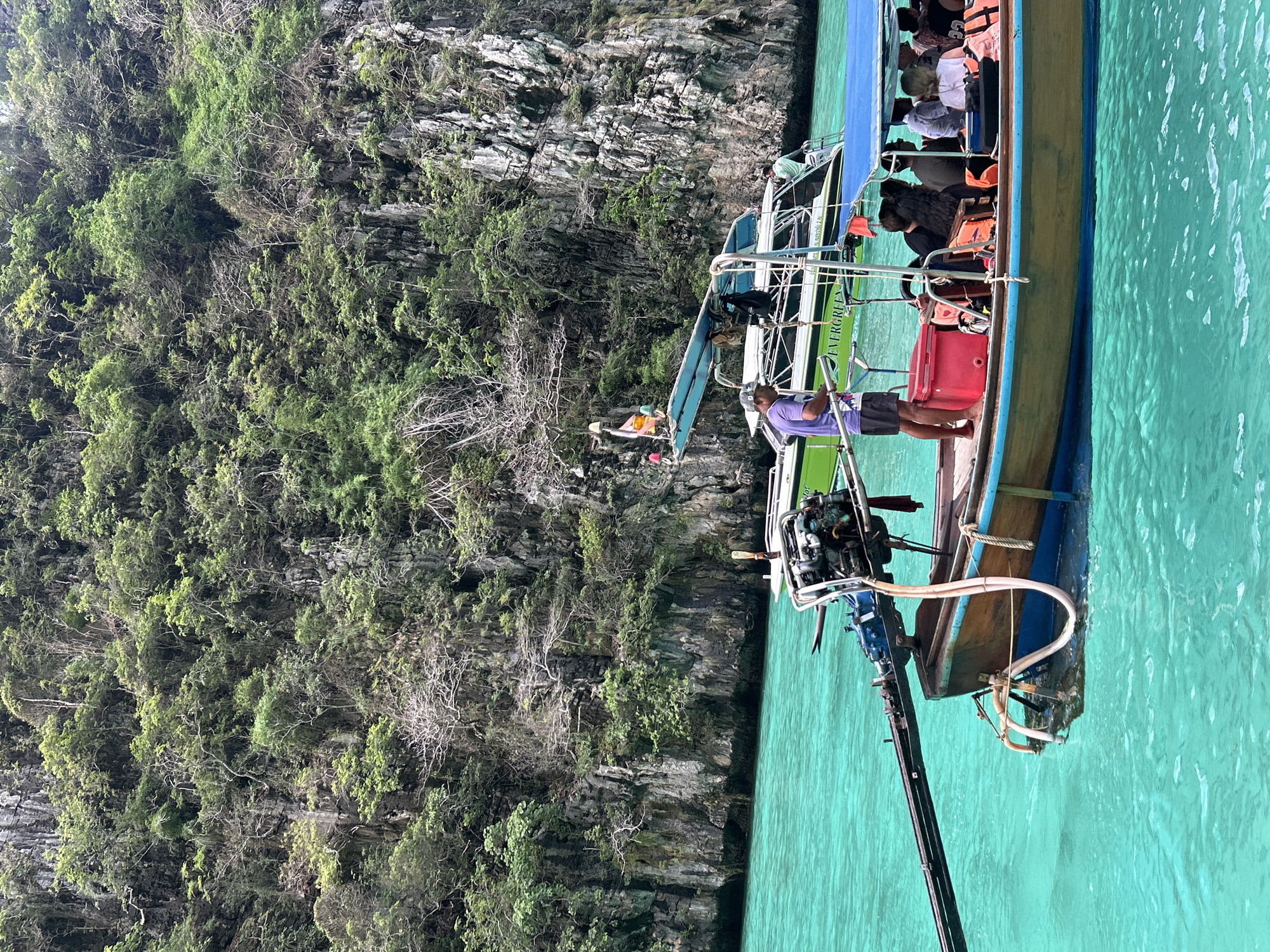 The image depicts a longtail boat in the turquoise waters of Ao Nang, Thailand, with a steep, jungle-covered limestone cliff in the background. The boat is wooden with a blue canopy and is carrying several passengers wearing life vests. A man in a purple shirt stands at the front of the boat, steering. Another boat, painted green and white with the word "EVERGREEN" visible, is anchored nearby. The scene suggests a typical tourist activity in the region, likely a boat tour of the islands.