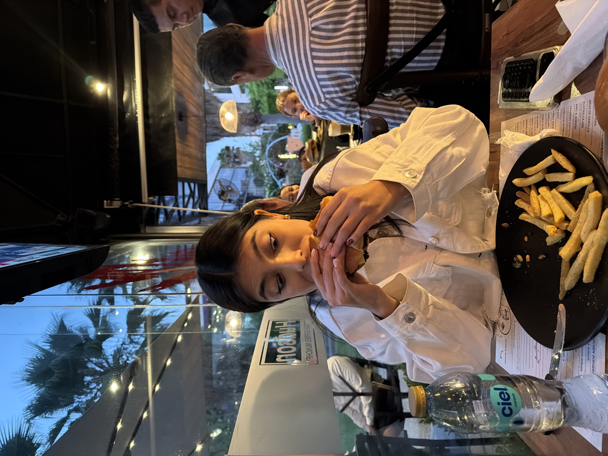 The image shows a woman in a white jacket eating a burger at a restaurant. A plate of fries sits next to her. A water bottle is on the table. Other people are sitting nearby, including a man in a striped shirt. The restaurant appears to have large windows that offer a view of palm trees. The OCR text on the image suggests that the location is in Mexico, with menu items listed in both English and Spanish. Based on the OCR text, the restaurant may be named "Haorou." It is impossible to pinpoint the exact location as San Bernardino Tlaxcalancingo from the image alone.