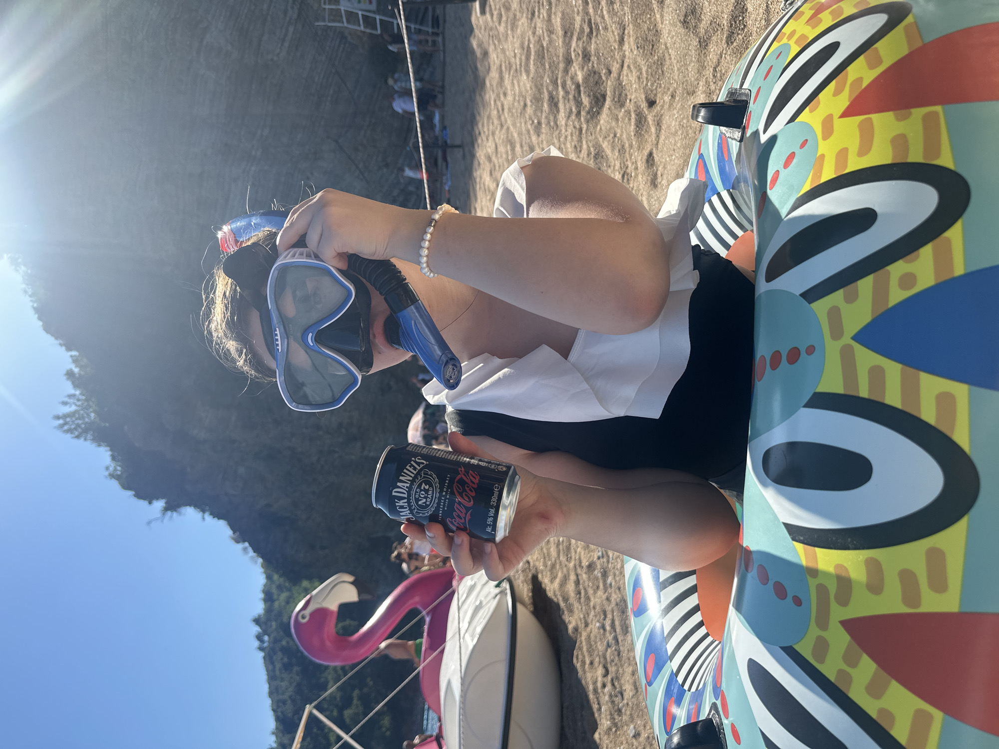 The image captures a person wearing a snorkel and holding a can of Jack Daniel's and Coca-Cola. The individual is wearing a white blouse and black pants. A pearl bracelet adorns their left wrist. They are sitting inside an inflatable ring with a colorful abstract design. The background suggests a beach setting with sand and what appears to be a coastal mountain range. The photo was taken outdoors under bright sunlight. Also visible are a white beach chair with a pink flamingo attached and other chairs on the sand. Based on the scenery and known travel destinations, this scene could potentially be located on a beach in or near Budva, Montenegro.