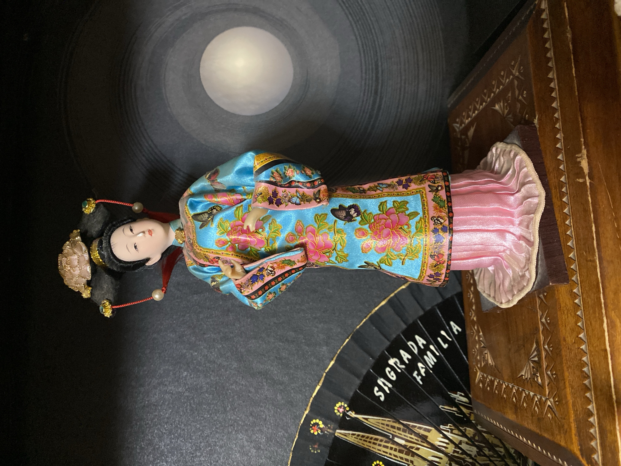 A doll representing a Chinese woman in traditional attire stands on a wooden box. The doll wears a brightly colored silk robe with floral and butterfly patterns, and a pink skirt. Her hair is styled in an updo with ornate accessories. To the left of the doll is a black fan with a depiction of the Sagrada Familia. The background appears to be a dark surface with a circular pattern and a lit orb. The wooden box has intricate carvings. There is no indication of a location in Novaya Usman', Russia.