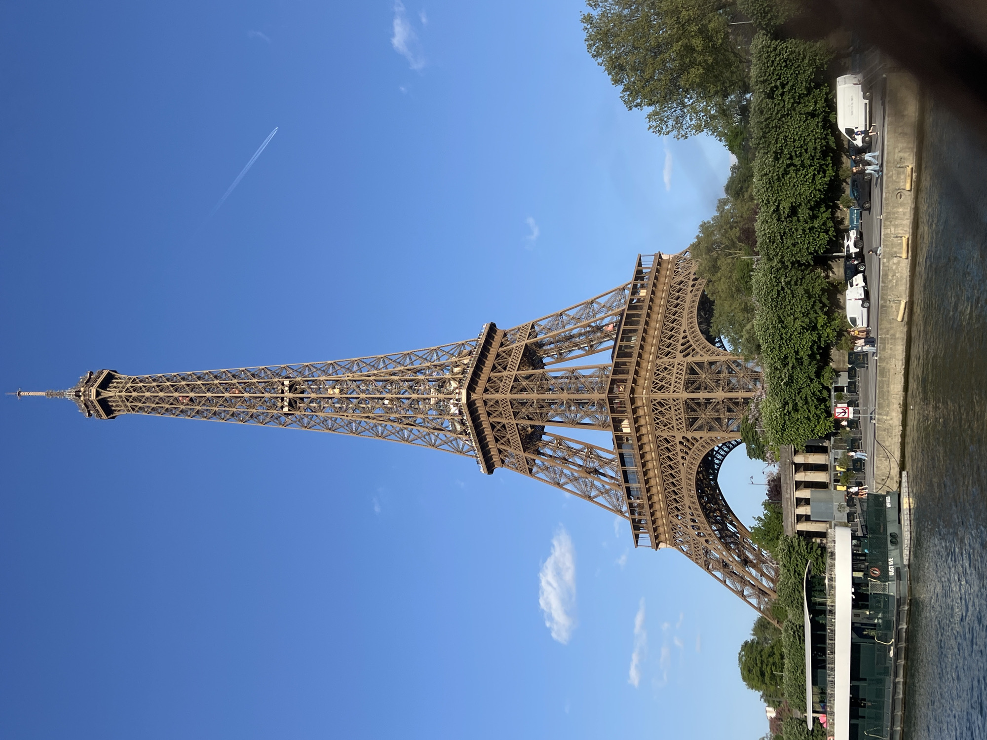The image captures a vibrant daytime scene in Paris, France, featuring the iconic Eiffel Tower prominently on the left side of the frame. The tower, viewed from a slightly low angle, showcases its intricate iron lattice structure against a clear, bright blue sky. A single, distinct airplane contrail streaks across the upper-left sky, accompanied by a few wispy white clouds.
To the right of the Eiffel Tower, the calm waters of the Seine River are visible, with a large, dark-hulled boat or barge docked alongside the bank. Green trees and dense bushes line the riverbank, creating a natural barrier between the river and a paved road. Several vehicles, including multiple white commercial vans and at least one dark car, are parked along this road. Text on one of the white vans reads "CHRONOPOST."
Scattered along the riverbank and near the parked vehicles are a number of people, some standing and others possibly seated, suggesting a mix of tourists and locals enjoying the riverside area. On the side of the boat, visible text includes "BATEAUX PARISIENS" and "QUAI J. CISSON," indicating it is a tourist vessel operating on the Seine.
The overall atmosphere suggests a pleasant, sunny day, ideal for sightseeing and outdoor activities in the heart of the city. The time of day appears to be mid-morning or afternoon, with ample natural light illuminating the scene.
