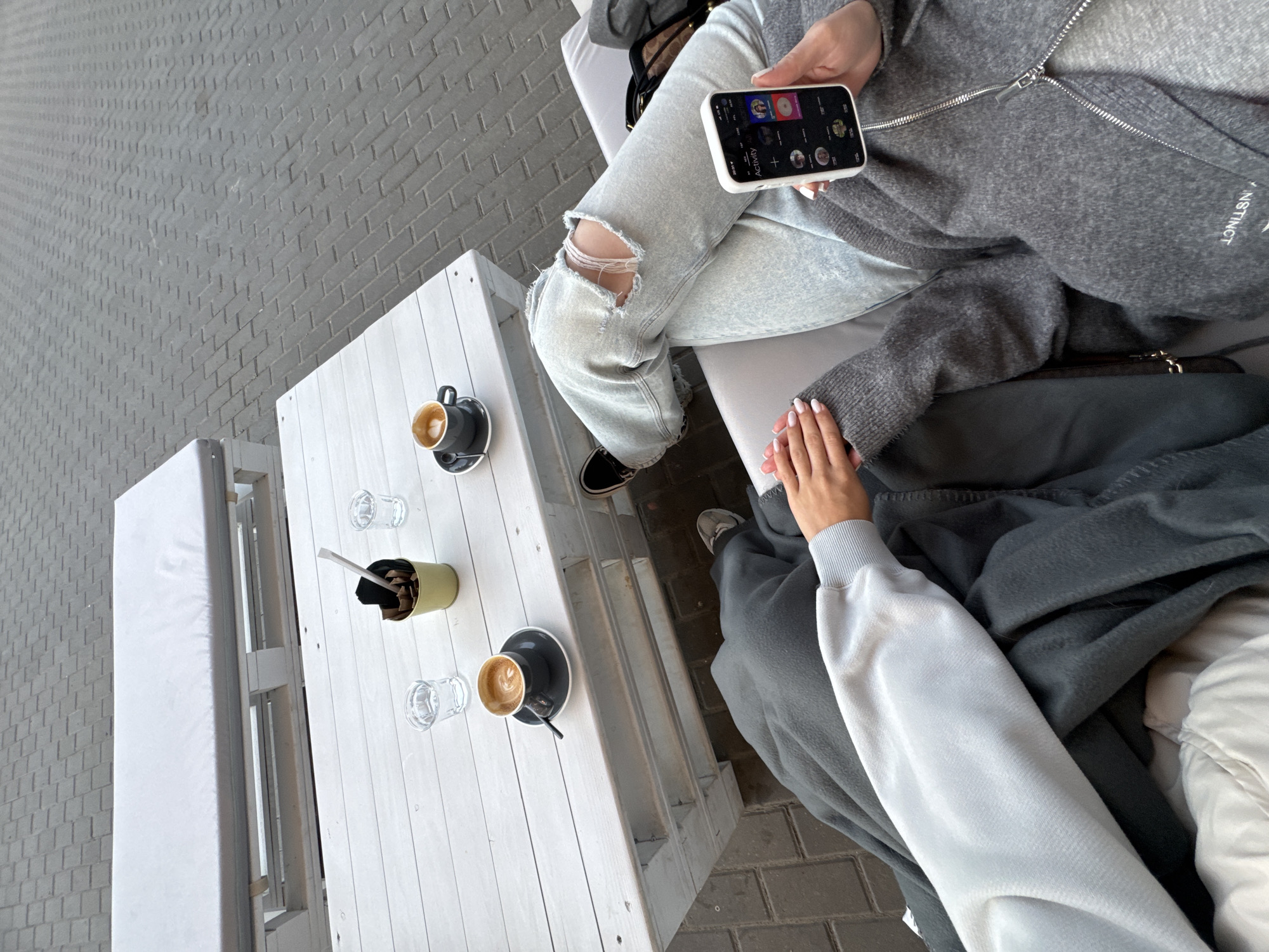 The image provides an overhead perspective of two individuals seated at an outdoor café or patio. One person, positioned to the left, is wearing light-wash ripped jeans, black sneakers, and a gray zippered cardigan. This individual is holding a white-cased smartphone with a screen displaying various app icons and partially visible text, including the word "Activity." Part of a white embroidered word, possibly "INSTINCT," is visible on their cardigan.
The other person, to the right, is wearing a light-colored long-sleeved top and is partially covered by a dark gray blanket or coat. Their hand is gently holding the hand of the person with the phone, suggesting a close and comforting interaction.
A white wooden slatted table, possibly constructed from pallets, sits between them. On the table are two cups of coffee, one featuring distinct latte art, two empty clear glasses, and a small green cylindrical container holding straws and possibly sugar packets. The floor is paved with gray rectangular bricks or pavers, common in outdoor settings. The bright, natural light indicates it is daytime. The scene conveys a relaxed and intimate social moment, possibly a casual coffee break between friends or partners.