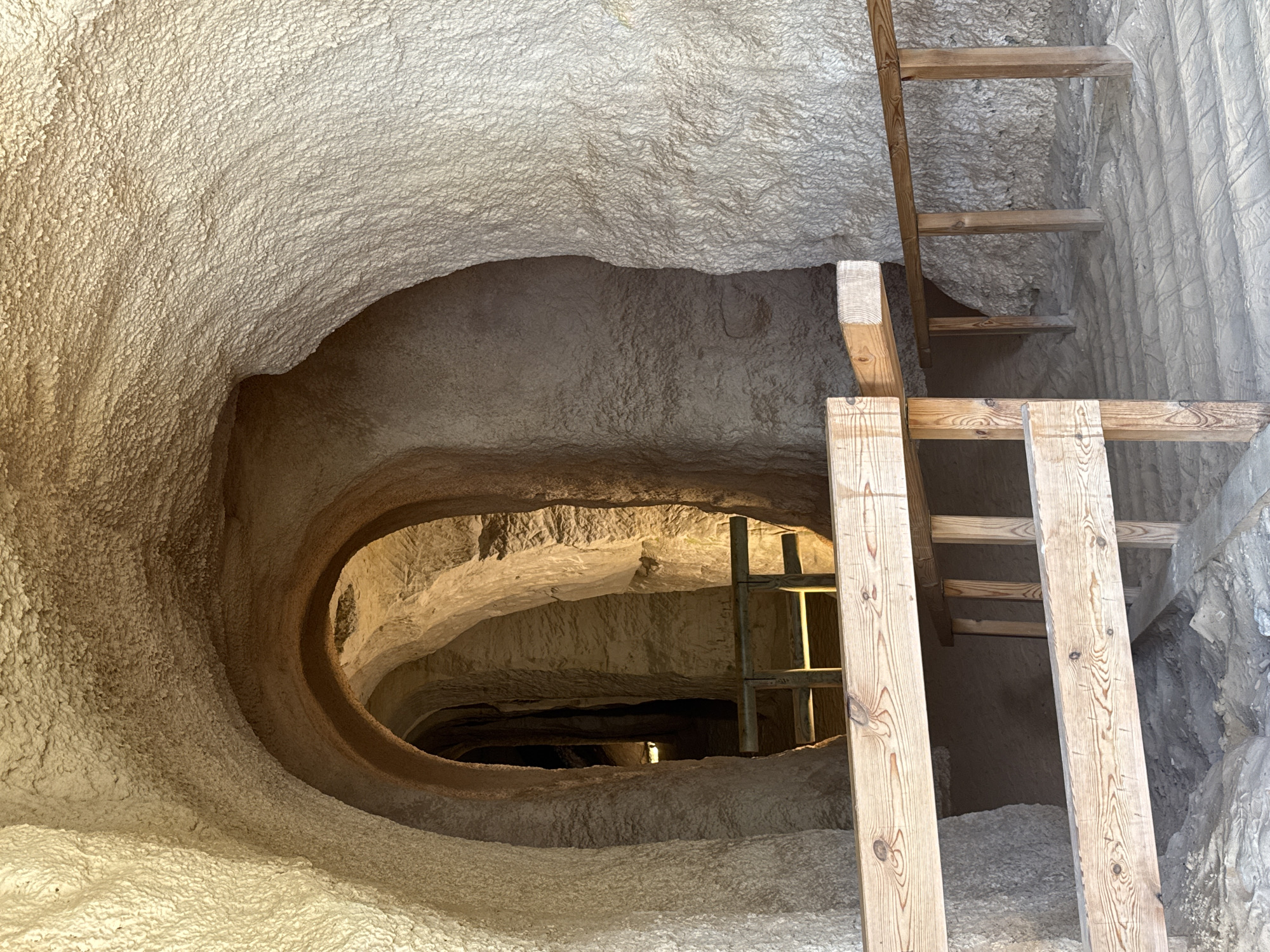 The image depicts the interior of the Piusa Sand Caves in Estonia. The scene focuses on a section of the cave system with arched passageways and sandy floors. Two wooden guardrails are prominently featured in the foreground, made of rough-hewn timber, with visible knots and wood grain. These rails guide visitors along a sandy path that is marked with parallel indentations.
The cave walls are rough and textured, appearing to be composed of compressed sand. The light source seems to be natural sunlight entering from an unseen opening, casting shadows and illuminating the textures of the cave. In the background, another arched opening leads to a darker, narrower passage, where a faint light is visible further within. The overall impression is one of a natural, yet managed, underground environment. There are no people visible in the image.