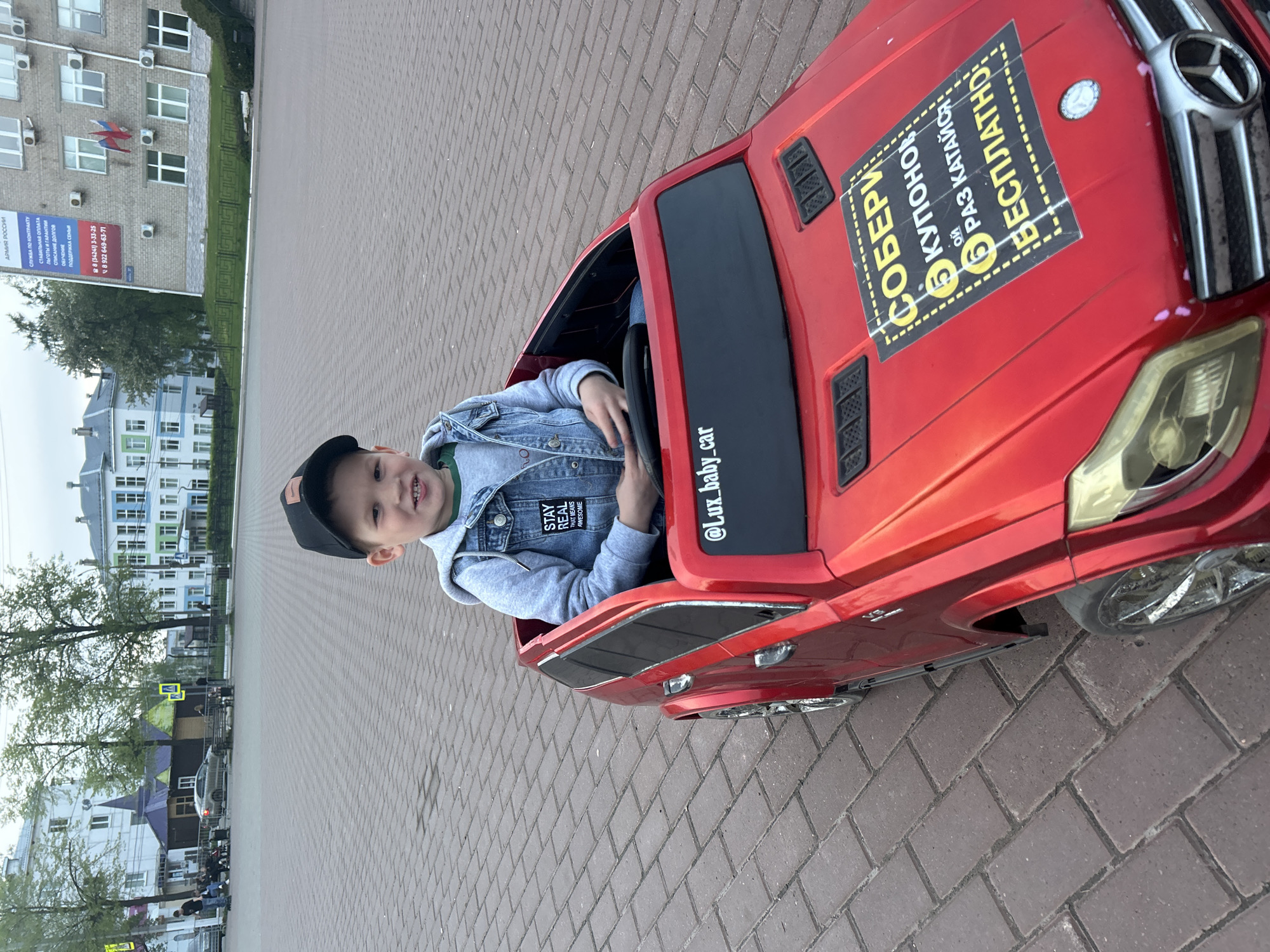 A young boy in a baseball cap and denim jacket smiles from inside a red Mercedes-Benz child's car, likely an electric toy. The car has a sticker on the hood advertising 5 free rides. The location is an outdoor brick-paved area, potentially a public square, in the city of Chaikovsky, Russia. Buildings are visible in the background, including one with a sign for the "Army of Russia" offering contract service, debt relief, and family support, along with a phone number. Trees and street signs are also present.