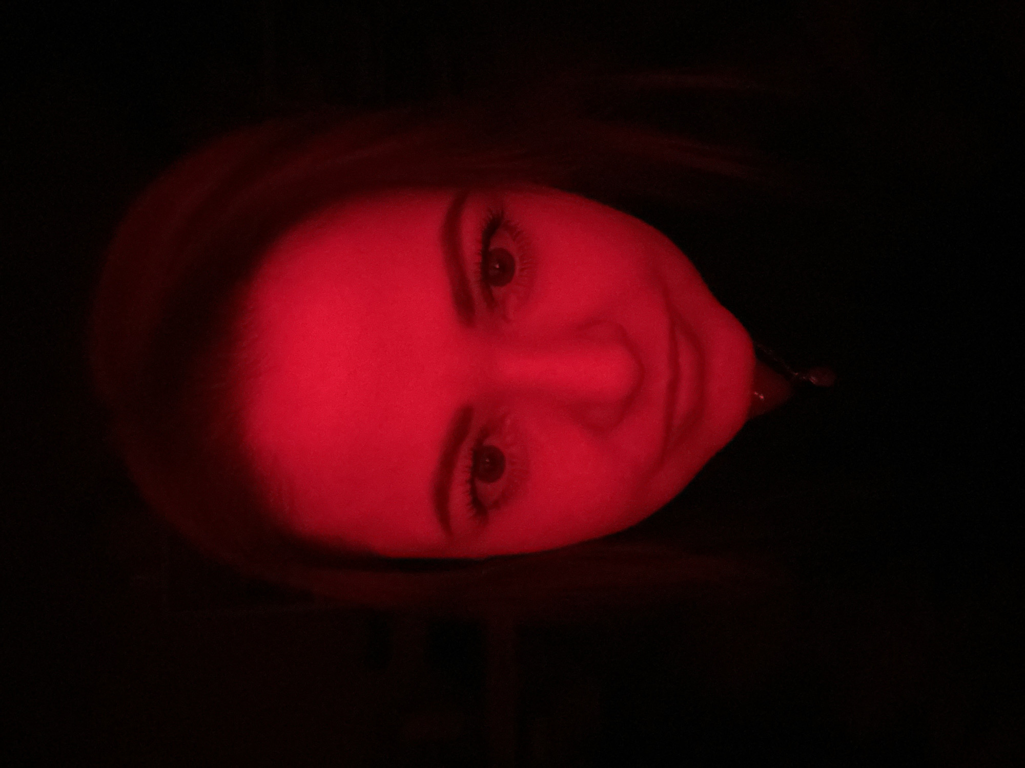 This close-up portrait captures a person's face, likely female, tilted horizontally, against a completely dark background. The face is dramatically illuminated by an intense red light, primarily from the left side of the frame, highlighting features such as the forehead, nose, cheeks, and chin. The subject's eyes are open and directed towards the viewer, with visible eyelashes and dark pupils. A subtle, soft smile is present on their lips, conveying a calm or slightly pleased expression.
The rest of the scene, including the person's hair and any surrounding environment, is submerged in deep shadow, emphasizing the isolated and illuminated face. This strong red light creates a striking, almost ethereal, and mysterious atmosphere, abstracting the setting. There are no other discernible objects or background details. The image suggests an indoor or controlled lighting environment, possibly at night, in Yekaterinburg, Russia. No text is visible in the media file.