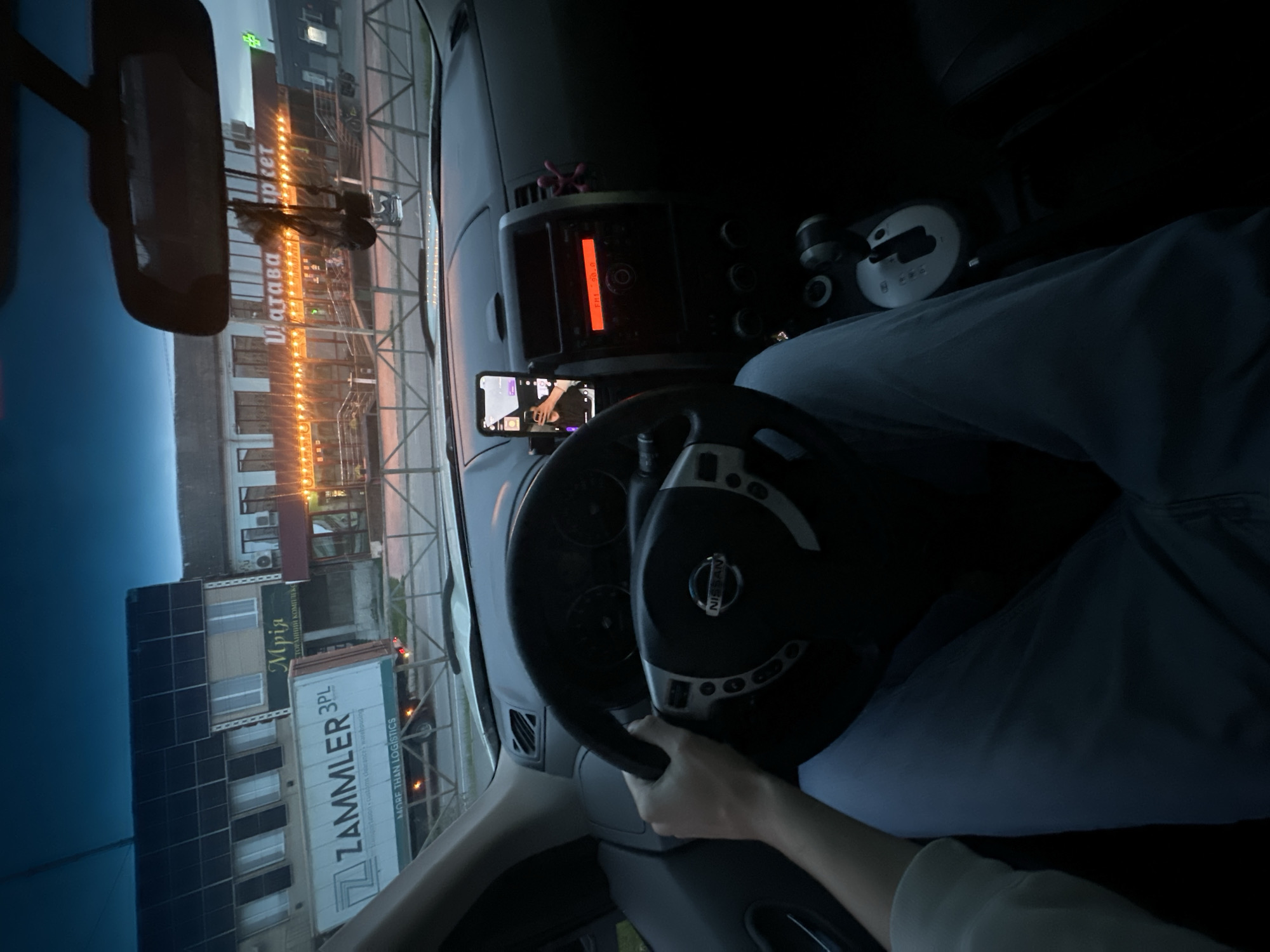 The image, taken from inside a Nissan vehicle, shows a person's hands on the steering wheel and their legs in light blue jeans. The car's dashboard is visible, with a phone mounted on the windshield displaying a selfie. Outside the car, across a chain-link fence, are buildings and a Zammler 3PL truck. In the background, a building with illuminated lights and signage that reads "Магава Ріет" (Matarva Market) is visible. The sky is dark, indicating it is either dusk or nighttime. Based on the signage, the location is identified as Shatava, Ukraine.