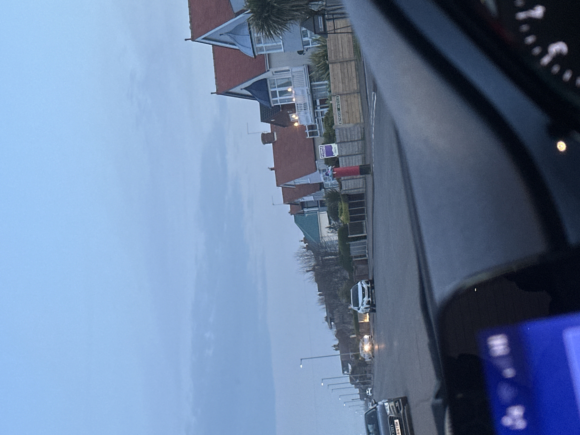The image, taken from inside a vehicle, shows a residential street in Gorleston-on-Sea, United Kingdom. Houses line the street, displaying varying architectural styles and colours. A red post box is visible near the houses. Several cars are parked or driving along the road. Street lamps are evenly spaced along the street. The sky is overcast and grey. The interior of the car, including the dashboard and digital display, is partially visible, framing the scene.