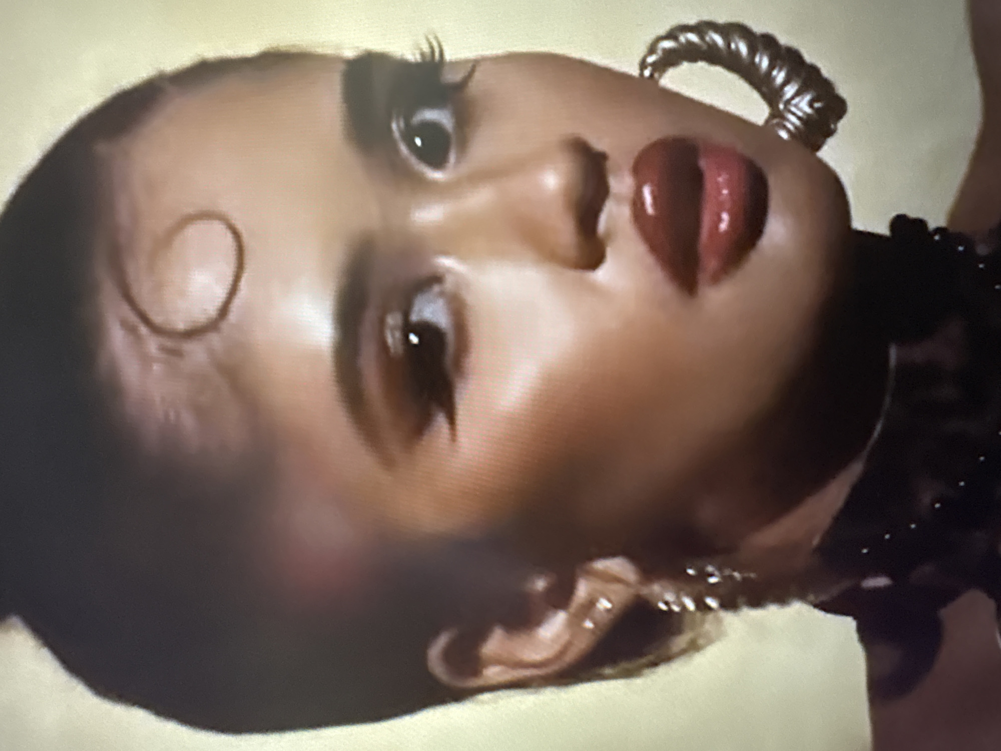This is a close-up, portrait-style photograph of a young woman. She has dark hair pulled back into a high bun, with a single curled strand of hair at her forehead. Her eyes are made up with dramatic, winged eyeliner and long lashes. She is wearing large, textured hoop earrings and a dark, possibly leather, choker with studs. Her lips are painted a glossy, deep red. The background is a muted, yellowish-gold color, suggesting an indoor studio setting or a plain wall. There is no indication of the city of Sofia, Bulgaria, or any other specific location.