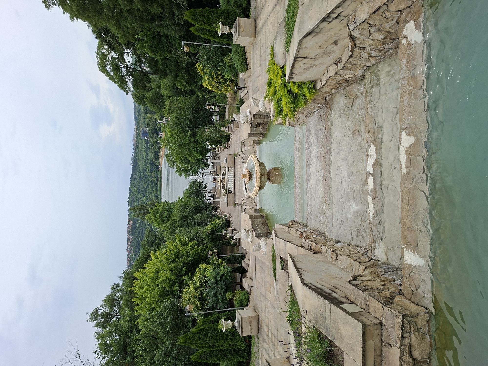 The image shows a multi-tiered fountain and staircase leading down towards a lake or reservoir, likely in the city of Chișinău, Moldova. The fountain features ornate basins and sculptures, including two fish-like spouts. The steps are made of stone and concrete, with some overgrown greenery. The surrounding area is landscaped with trees, shrubs, and decorative urns. In the distance, a body of water is visible, bordered by a tree-covered hillside and a town. The sky is overcast. The scene depicts a public park or recreational area.