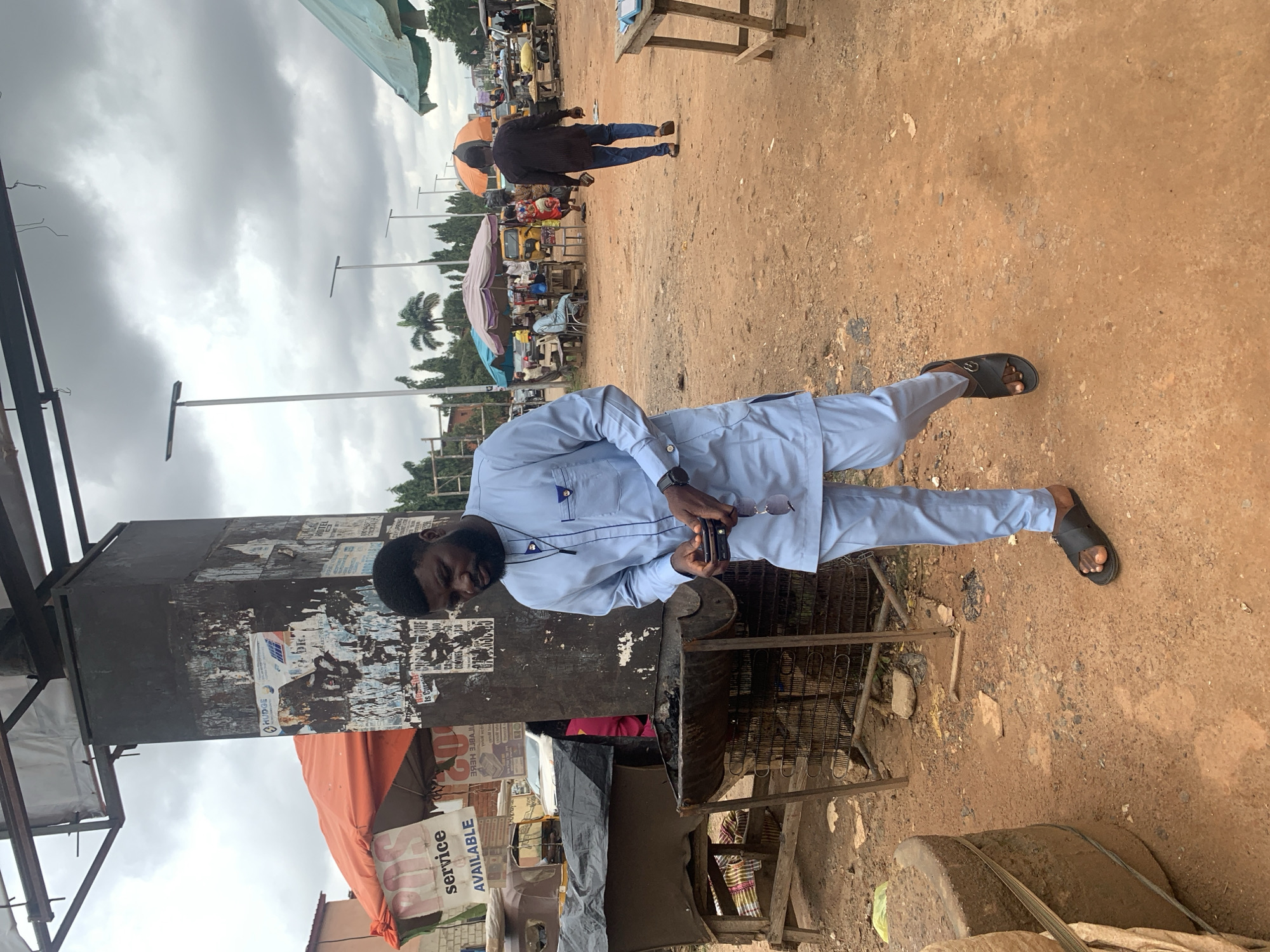 A man wearing a light blue traditional Nigerian outfit with matching trousers and sandals walks through an outdoor market in Abuja, Nigeria. He is holding a phone in his right hand and sunglasses in his left hand. The market is dusty and appears to be under a cloudy sky. There are stalls with umbrellas, vehicles, and other people in the background. A large, dark pillar with posters is to the left of the man, and a metal grill structure is in front of him.
