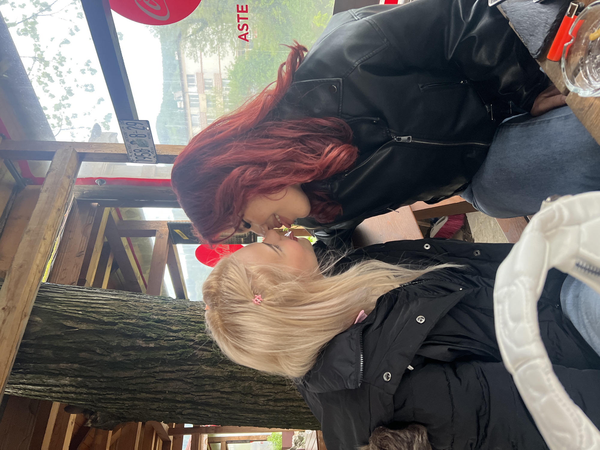 Two women are facing each other. The woman on the left has strawberry blonde hair, with a light black jacket with a furry lining on the hood. The woman on the right has red hair and is wearing a black leather jacket with blue jeans. They are indoors, possibly in a restaurant or cafe, with wooden beams visible in the background. In the background is a window with an ad for "ASTE." Through the window, there are buildings with foliage. Additionally, there is a decorative license plate, with "MICHAEL JACKSON" and "1958-8-29" on it. There is also a wooden table.