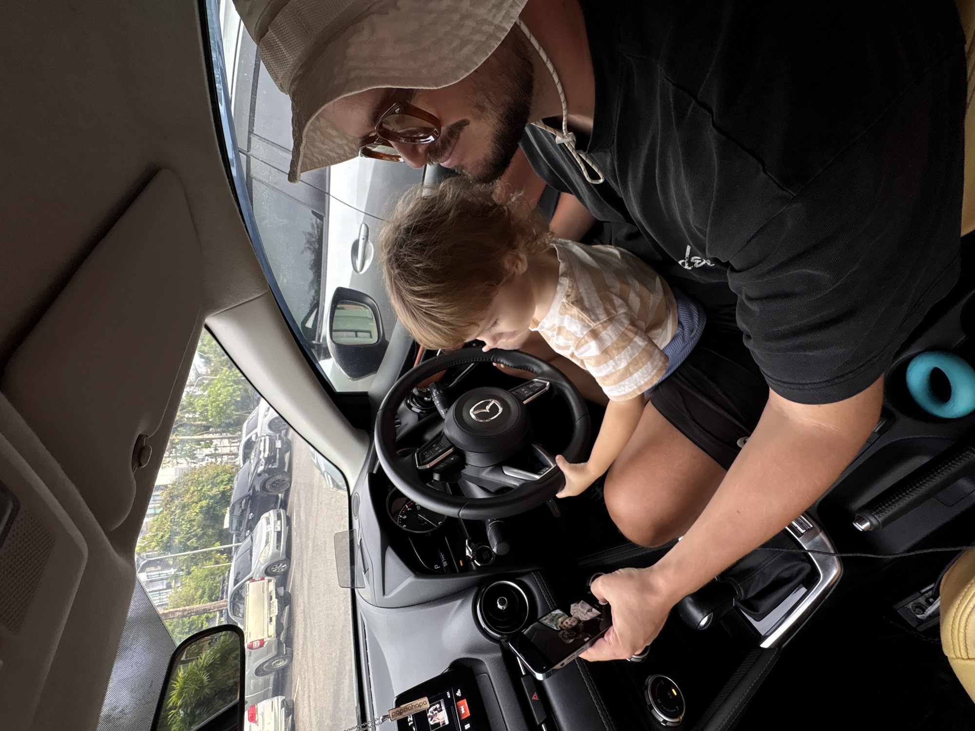 An adult male wearing sunglasses and a bucket hat, with a beard, is sitting in the driver's seat of a parked car. He is holding a smartphone in his left hand, displaying a picture of himself and a child. Next to him, on his lap, is a young child. The child's hands are on the steering wheel, appearing to mimic driving. The car's interior is visible, including the dashboard, steering wheel, gear shift, and a heads-up display. Outside the car, other vehicles are parked in what appears to be a parking lot. The environment suggests an outdoor setting, likely in a city, as indicated by the presence of parked cars and buildings in the background. The scene is set in Choeng Thale, Thailand.