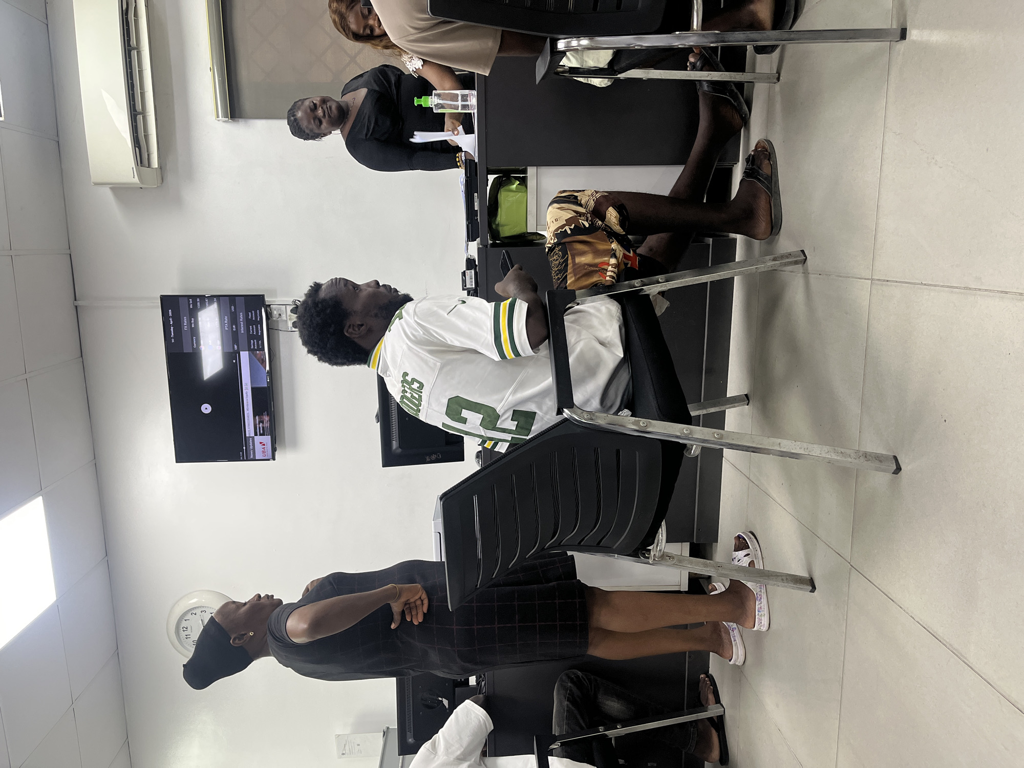 The scene is an office space in what appears to be Ado, Nigeria, judging by the style of dress and the content on the television screen. There are several individuals present. A man wearing a Green Bay Packers jersey is seated in a black office chair, looking towards a television mounted on the wall. The television is displaying what seems to be financial market rates. A woman dressed in black stands next to the man, her arms crossed. Another woman, also in black, is sitting at a desk behind them. Another person is seated at the desk, wearing beige. A white wall-mounted clock is visible. The office appears to be air-conditioned.