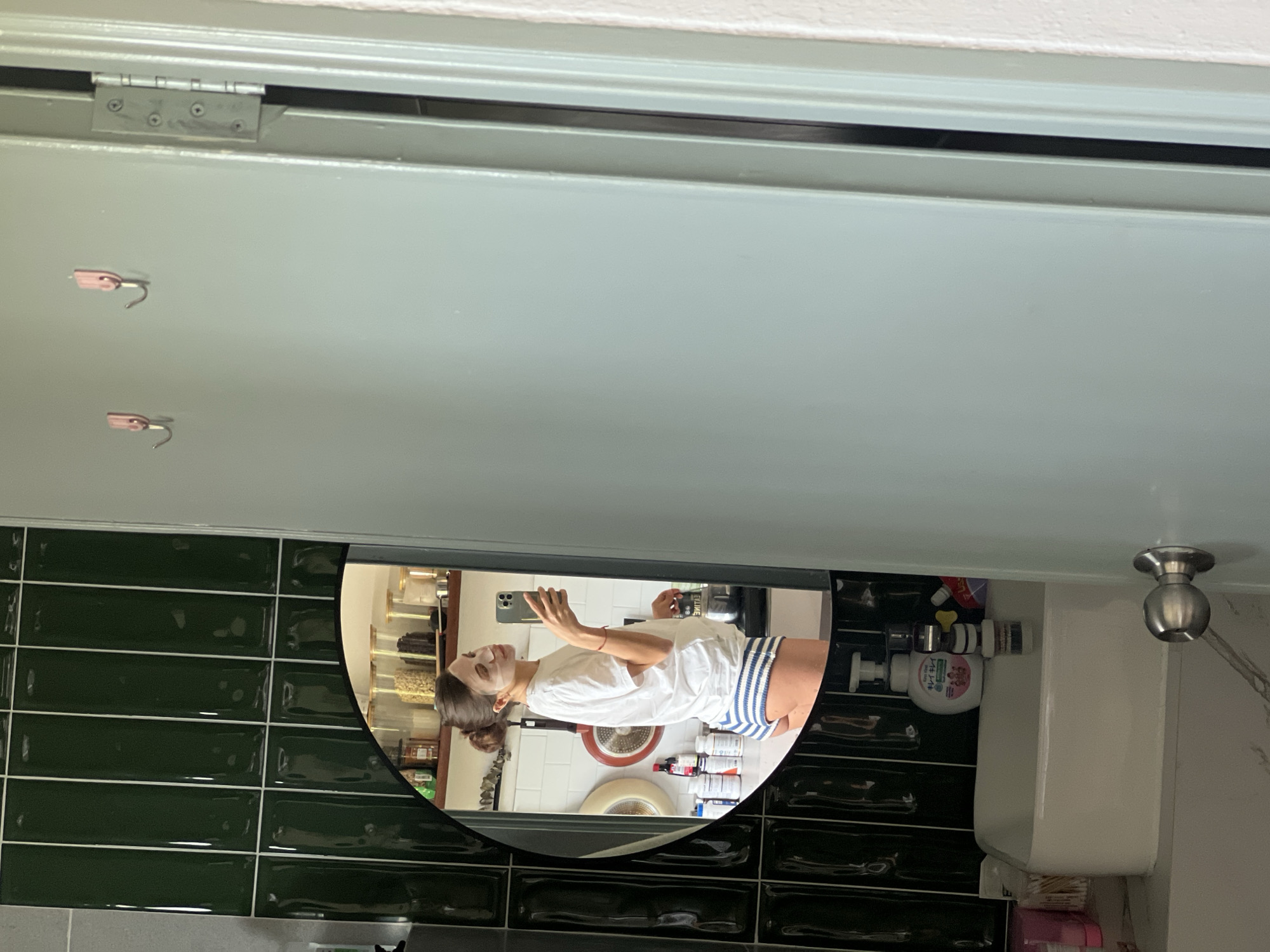 In a bathroom with dark green subway tile walls, a person with their hair in a bun is taking a mirror selfie. They are wearing a white t-shirt and blue and white striped shorts, and have a face mask on. The mirror reflects shelves in the background with jars of food items and bottles. A white sink is visible in the foreground, with various toiletries on it. The scene is likely in a residential bathroom in Ko Samui District, Thailand.