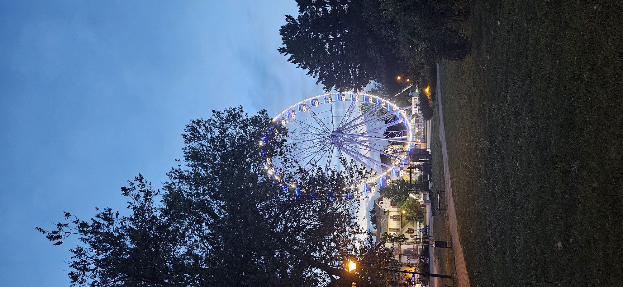The image depicts a Ferris wheel illuminated with blue and white lights, located in the city of Keszthely, Hungary. The Ferris wheel is situated in a park-like setting with trees and grassy areas. In the background, there are buildings that appear to be part of the Balaton region. The sky is a deep blue, indicating it is either dusk or dawn. A lamppost with an orange glow stands to the left of the Ferris wheel, and a bench is visible on a paved path. The scene is peaceful and serene, with no people clearly visible in the foreground.