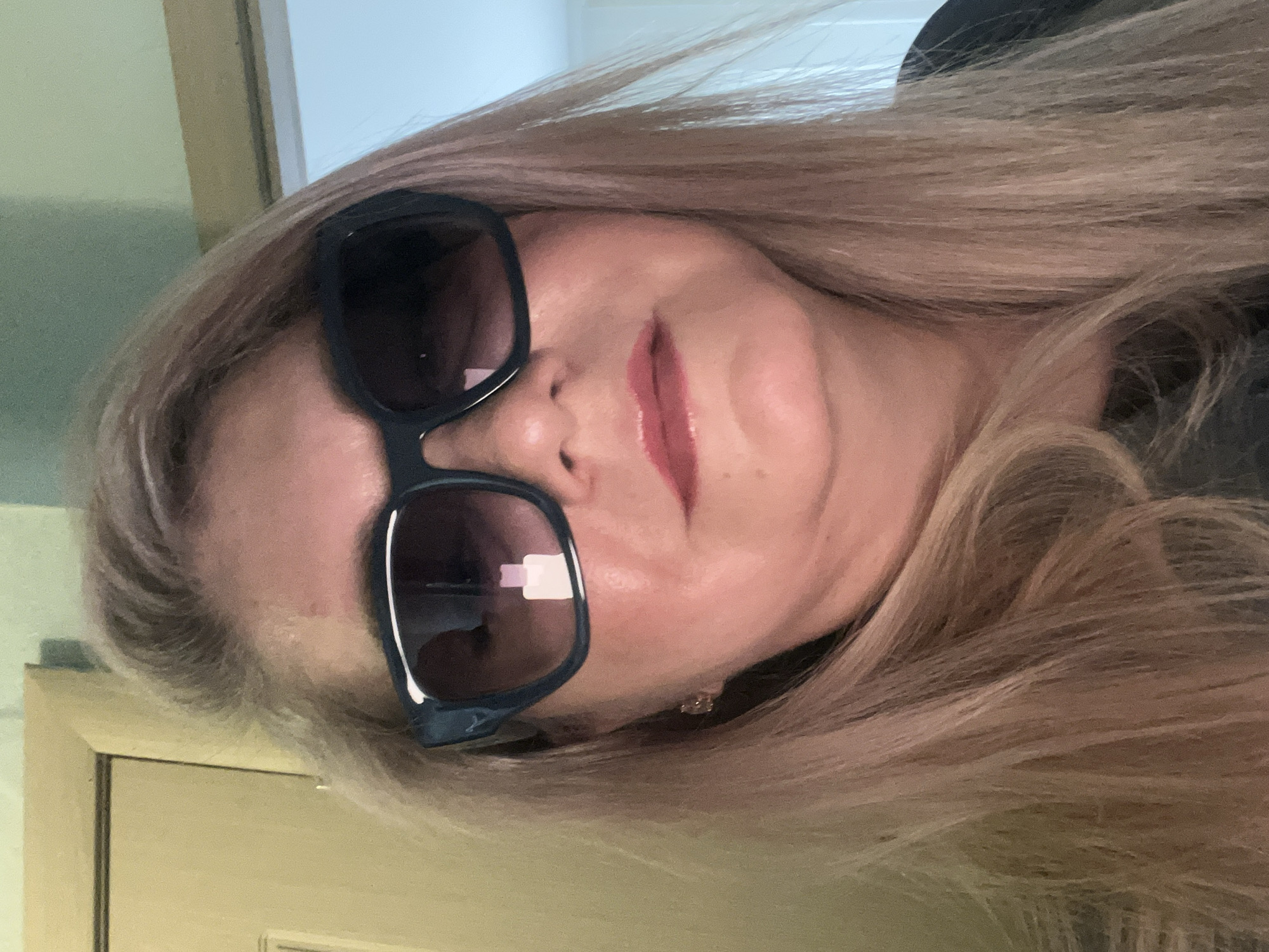 A woman with fair skin and long, light blonde hair poses for a close-up photograph while wearing dark sunglasses. Her lips are coated in a pink lipstick. A dark-colored garment is visible around her neck. A door or wall is partially visible in the background, suggesting an indoor setting. The image contains no information to identify the location as Izhevsk, Russia, or any other city.