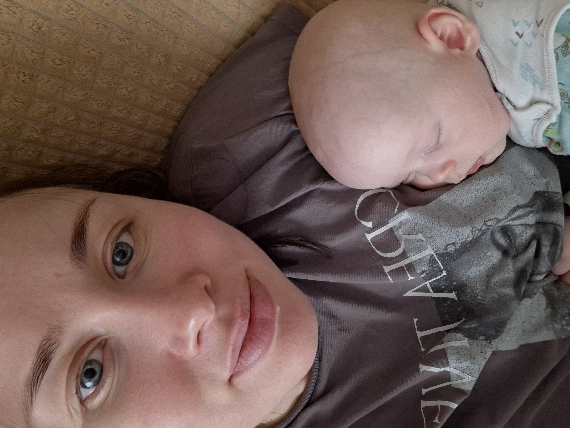 A close-up, high-angle shot captures a woman and a sleeping baby. The woman is positioned on the left side of the frame, lying down and looking directly at the camera with clear blue eyes and neatly groomed dark eyebrows. Her expression is calm and gentle, with full lips slightly parted. She is wearing a dark grey t-shirt, on which large, distressed white letters are visible, spelling "CREATE". A faded graphic, possibly a face, is underneath the text on her shirt.
The baby is nestled on the woman's right shoulder/chest, fast asleep. Only the baby's head and a small portion of their body are visible. The baby has very fair skin and appears bald or with very fine hair. Their eyes are closed peacefully, and a small ear is visible. The baby is wearing a light-colored garment, possibly a onesie, with subtle patterns of what appear to be tiny white shapes against a pale background.
The background consists of a textured, light brown fabric, likely a couch cushion or blanket, suggesting an indoor setting. The lighting is soft and even, highlighting the subjects. The overall scene conveys a sense of intimacy, peace, and maternal bonding.