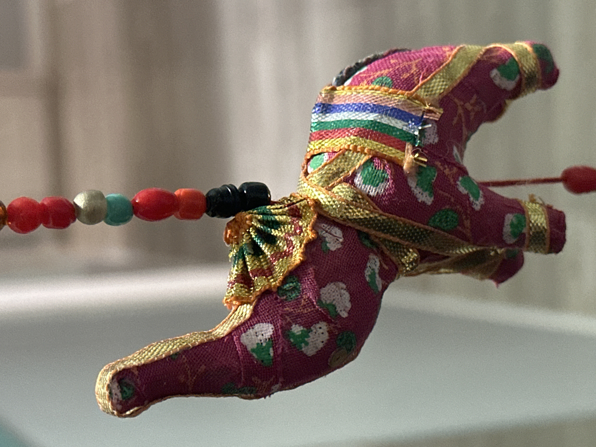 This is a close-up, eye-level shot of a decorative fabric elephant ornament. The elephant is primarily pink with a pattern of green and white floral shapes. It is adorned with gold trim and a colorful striped fabric draped over its back. A string of red, silver, turquoise, and black beads hangs above the elephant. The background is softly blurred, appearing to be an indoor setting with a light-colored wall and a hanging fabric. There is no visible indication of the city of Nevinnomyssk, Russia, in the image.