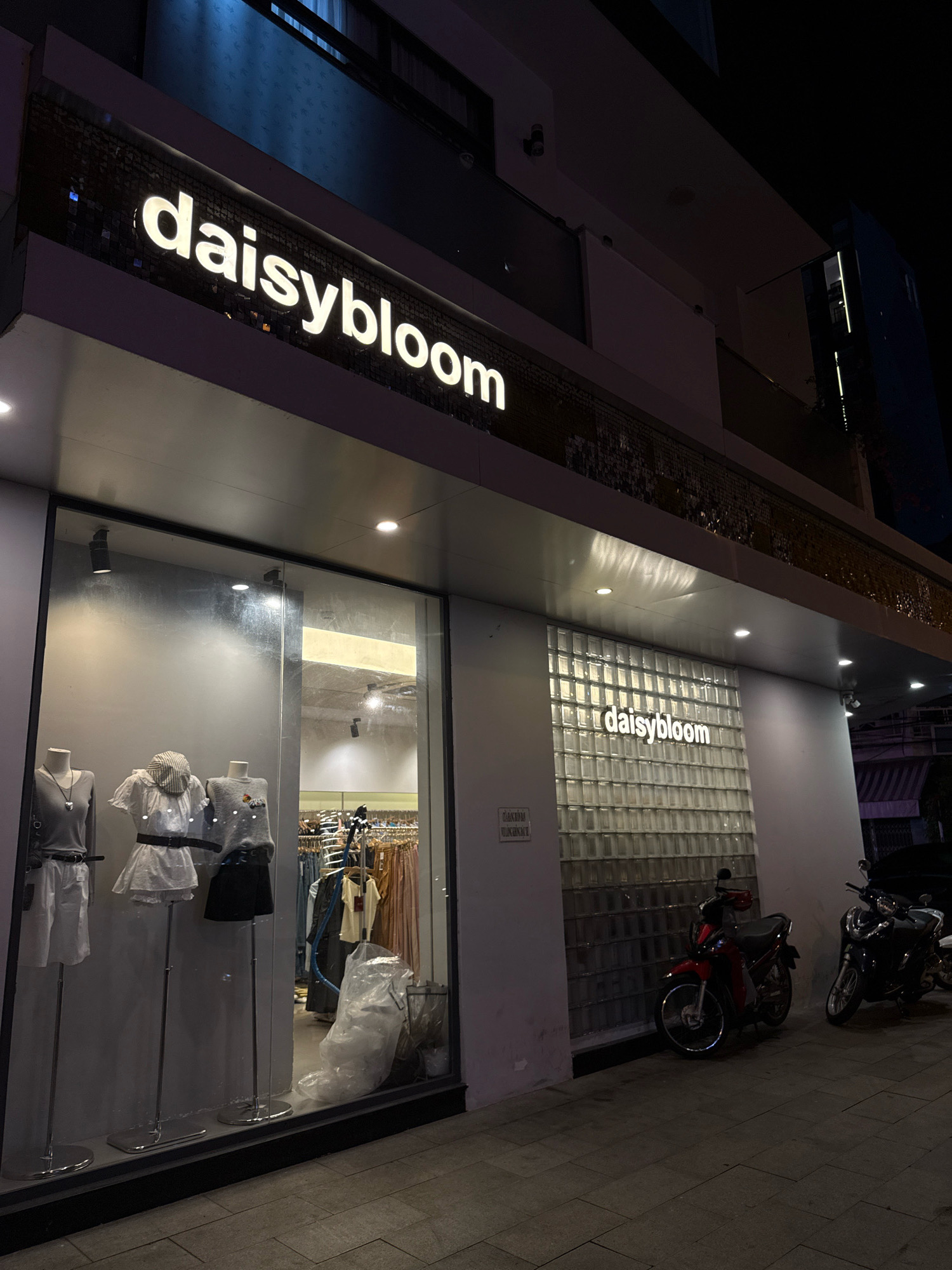 The image captures the exterior of a store named "daisybloom" at night in Nha Trang, Vietnam. The store has a prominent illuminated sign with white lettering that reads "daisybloom". The storefront features a large glass window displaying mannequins dressed in fashion items. To the right of the window is a wall made of glass blocks, also with an illuminated "daisybloom" sign. Two motorcycles are parked on the sidewalk in front of the store. The overall scene depicts a commercial establishment in an urban setting at night.