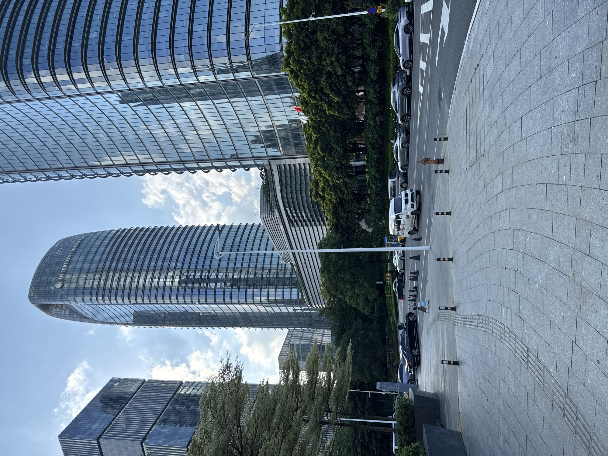 This is a street-level, eye-level, vertical shot of a modern urban environment in Haizhu District, China. The scene is dominated by towering glass skyscrapers reflecting the sky and clouds. In the foreground, a paved walkway with textured tactile paving for the visually impaired curves towards the street. Several cars are parked and driving on the street, including a white van, a black sedan, and lighter-colored sedans and SUVs. Traffic cones and bollards are visible along the roadside. Lush green trees line the street between the buildings and the sidewalk. The sky is partly cloudy, indicating daytime. The overall impression is of a well-developed business district.
