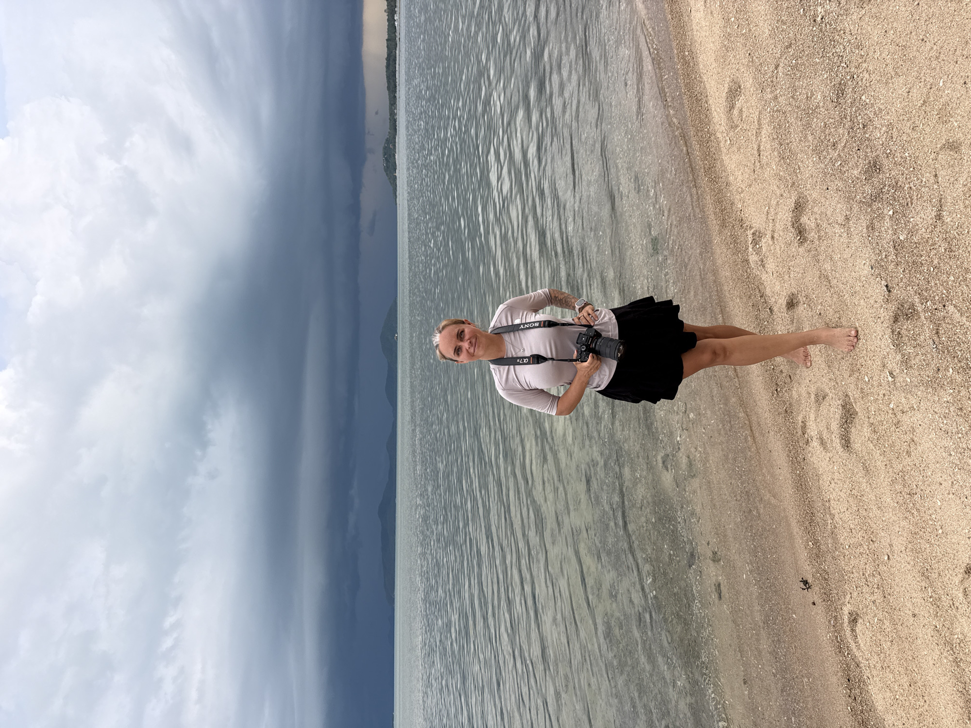 A medium shot captures a woman standing on a sandy beach, facing forward with a camera around her neck. She has light skin, blond hair pulled back, and is wearing a light-colored t-shirt and a black skirt. The camera strap is visible, with the camera body resting against her stomach. Her left hand is on her hip, and her right hand is near the camera. The background consists of the sea with gentle ripples and a cloudy, dark sky, suggesting an impending storm. In the distance, a hazy shoreline with hills is visible. The sand in the foreground shows footprints and a small crab. The location is identified as Ban Bang Khao, Thailand.