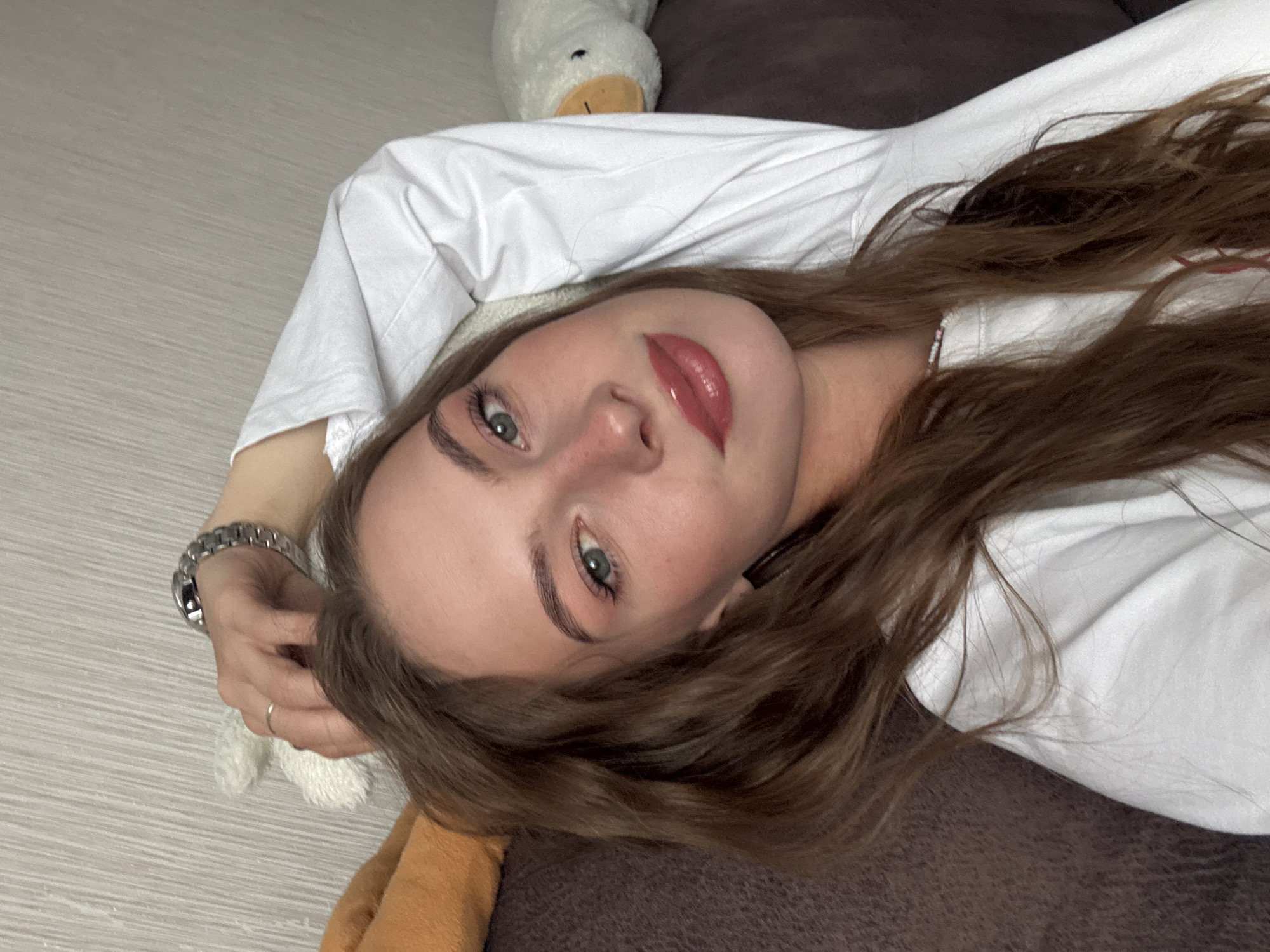 A young adult female is depicted in a reclining position, looking directly at the camera. She has long, wavy brown hair, fair skin, and blue-green eyes enhanced with mascara. Her cheeks and nose show a flush of pink blush, and her lips are covered in a glossy, light red or coral tint. She wears a plain white long-sleeved t-shirt. Her left arm is bent, with her hand resting near the top of her head; a silver-toned metal wristwatch with a link bracelet is visible on her left wrist, and a simple silver-toned band ring is on her left ring finger. She is situated on a dark brown upholstered surface, likely a sofa or bed. To her left, an orange-brown cushion and a small, white, fluffy object are visible. To her right, a white stuffed animal resembling a duck or goose with an orange beak rests on the same brown surface. The background consists of a light beige or grey wall featuring a subtle vertical textured pattern. The scene is indoors and evenly lit.