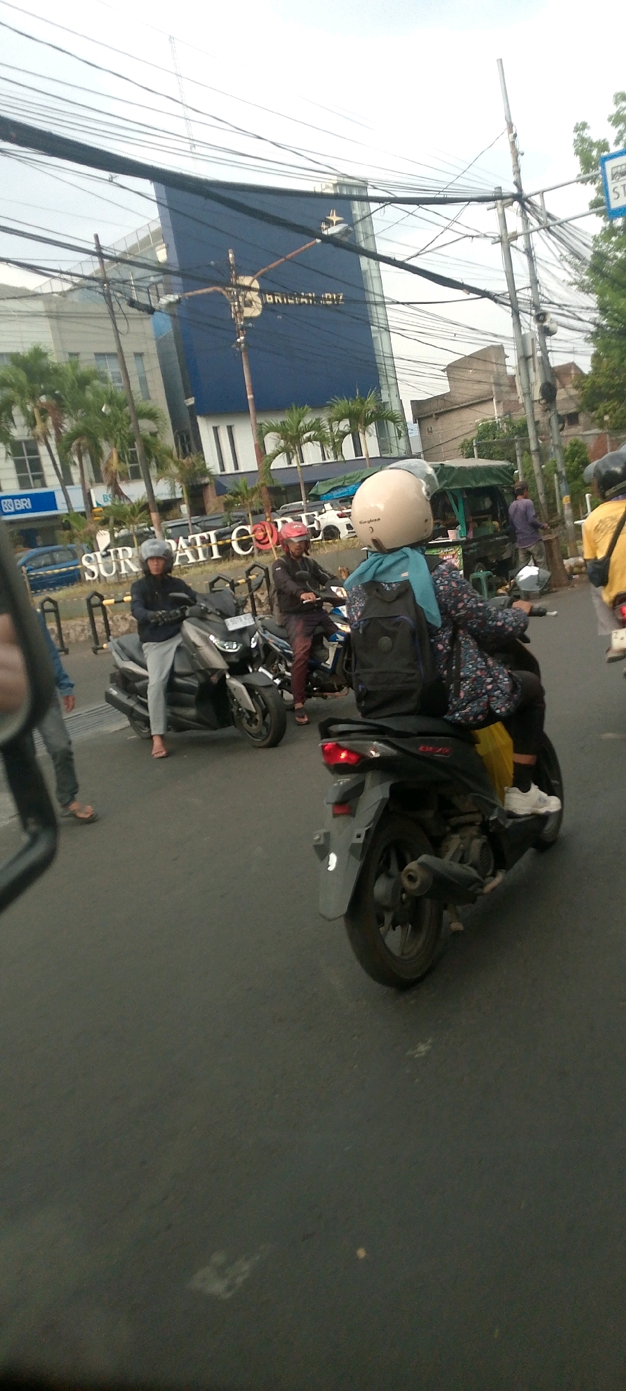 The image depicts a street scene in Bandung City, Indonesia, likely during daylight hours given the ambient light. Several individuals are on motorcycles, suggesting a traffic scenario. In the background, a modern blue building with the text "BRIGITANT BIZ" is visible, indicating a commercial area. Palm trees line the street, adding to the tropical urban landscape. The foreground shows the rear of a motorcycle, with its taillight illuminated, and parts of the road surface. Wires are strung across the sky, a common sight in Indonesian cities. The presence of multiple motorcycles and people suggests everyday urban activity.