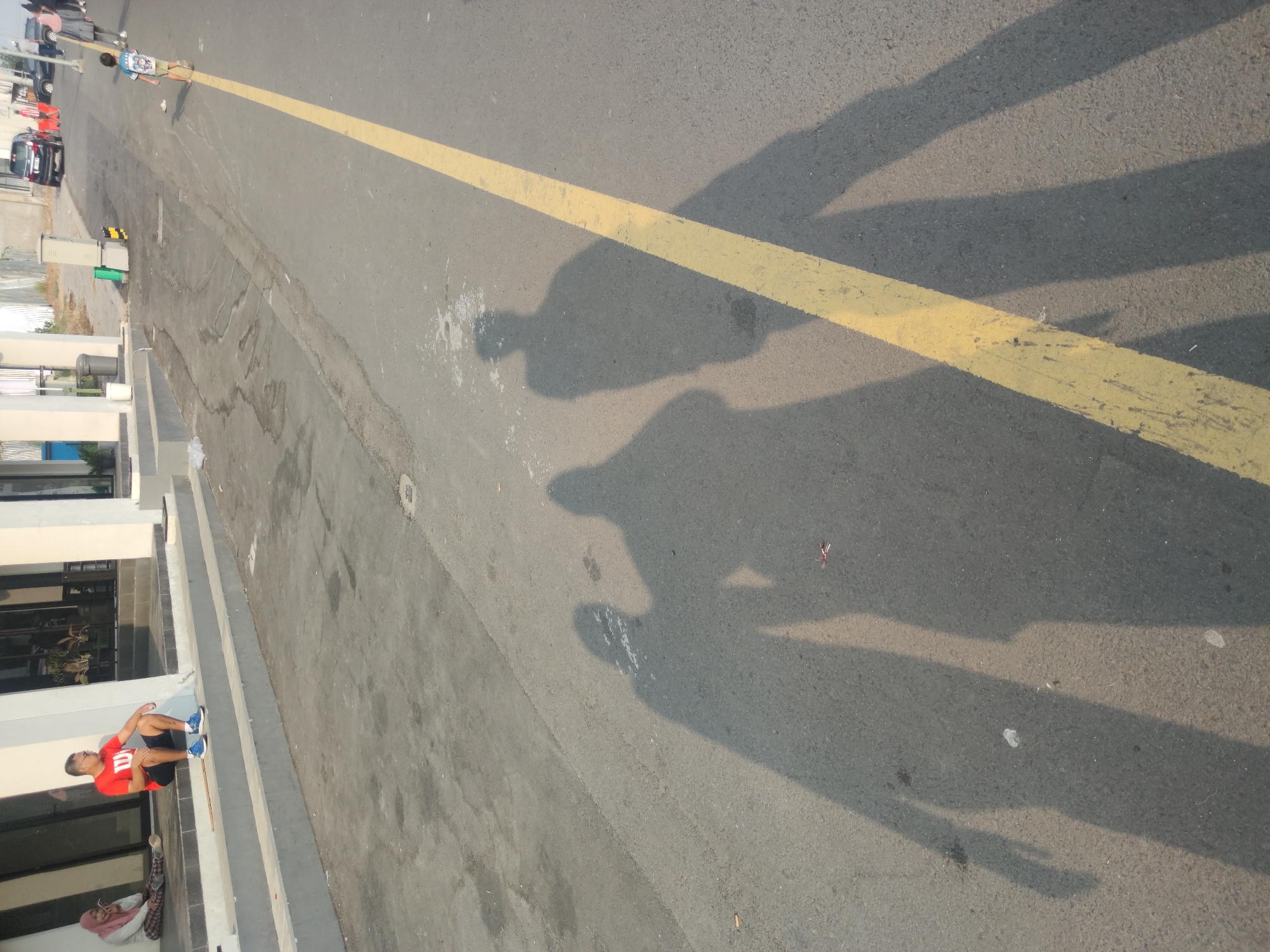 The image depicts an outdoor scene at the city of Petiken, Indonesia, under daylight. In the foreground, the long shadows of three individuals are cast across an asphalt road marked with a yellow line. The shadows suggest they are standing or walking away from the camera. The middle shadow appears to be that of a single person, while the shadows to its left and right are of at least two people, one of whom is taller. In the mid-ground, a young child in a t-shirt and shorts is walking away from the camera on the right side of the road, with other pedestrians and parked vehicles visible further in the distance on the right. The background features a building with an entrance, where two individuals are seated on steps. One is a person wearing a headscarf and the other is a man in a red t-shirt and black shorts. The environment is urban with paved surfaces and architectural structures.