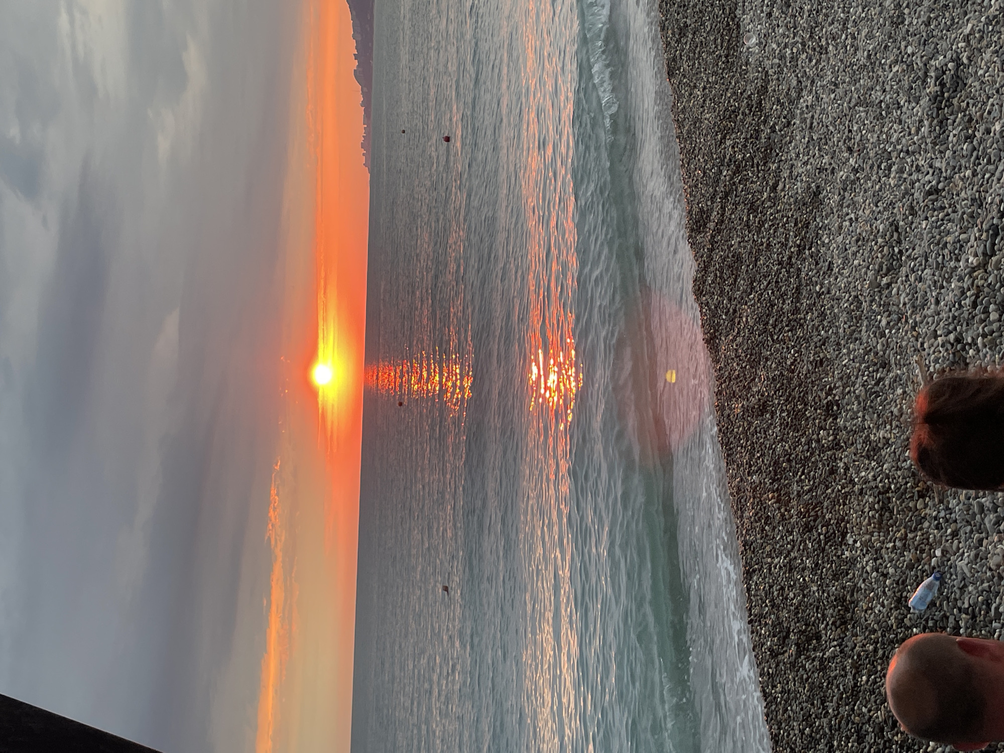 The image depicts a sunset scene at a pebble beach in Khostinskiy, Russia. The sun, a bright orange orb, is low on the horizon, casting a golden and orange glow across the sky and reflecting shimmering patterns on the surface of the calm sea. The sky is partly cloudy, with some darker clouds on the left side of the frame contrasting with the warm hues of the sunset.
The foreground is dominated by a pebbled beach, with small, dark grey stones covering the ground. The sea gently laps at the shore, with small waves creating a textured pattern on the water. In the distance, a faint silhouette of land or buildings can be seen along the horizon, hinting at a coastline.
The image appears to be taken from a low angle, looking out towards the sea and the setting sun. Two sets of human feet are visible in the bottom right corner, suggesting someone is standing or sitting on the beach, observing the sunset. A single plastic bottle lies on the pebbles near the water's edge, a small detail amidst the natural beauty of the scene. The overall mood is serene and peaceful, capturing the tranquil beauty of a sunset by the sea. The time of day is clearly sunset, and the weather appears calm with mild cloud cover.