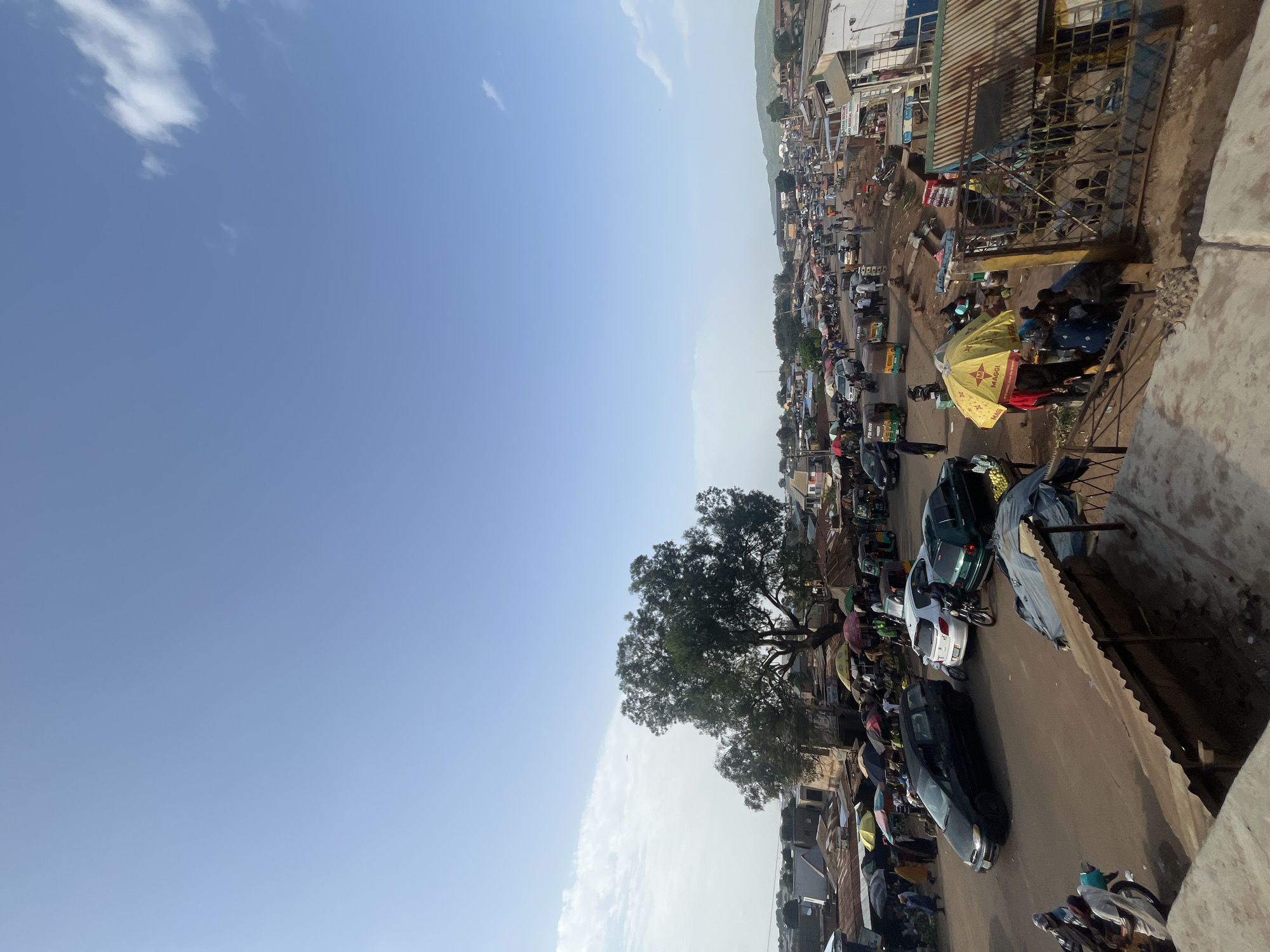 The image shows a bustling street scene in Rafin Par, Nigeria. The sky is clear blue with a few wispy clouds. In the foreground, there are several market stalls with corrugated metal roofs, some covered with tarps. A few people are gathered around these stalls.
Further down the street, there is a busy marketplace with many people and various vehicles. Cars, motorcycles, and what appear to be auto-rickshaws, known locally as Keke Napep, are navigating the unpaved road. Market stalls are set up along both sides of the road, with many vendors using umbrellas for shade. Some of the stalls are selling produce, and others appear to be general merchandise.
A large, mature tree stands prominently in the middle ground, casting a shadow over some of the market stalls. In the background, there are more buildings and a distant hilly landscape. The overall atmosphere suggests a vibrant and active commercial hub.