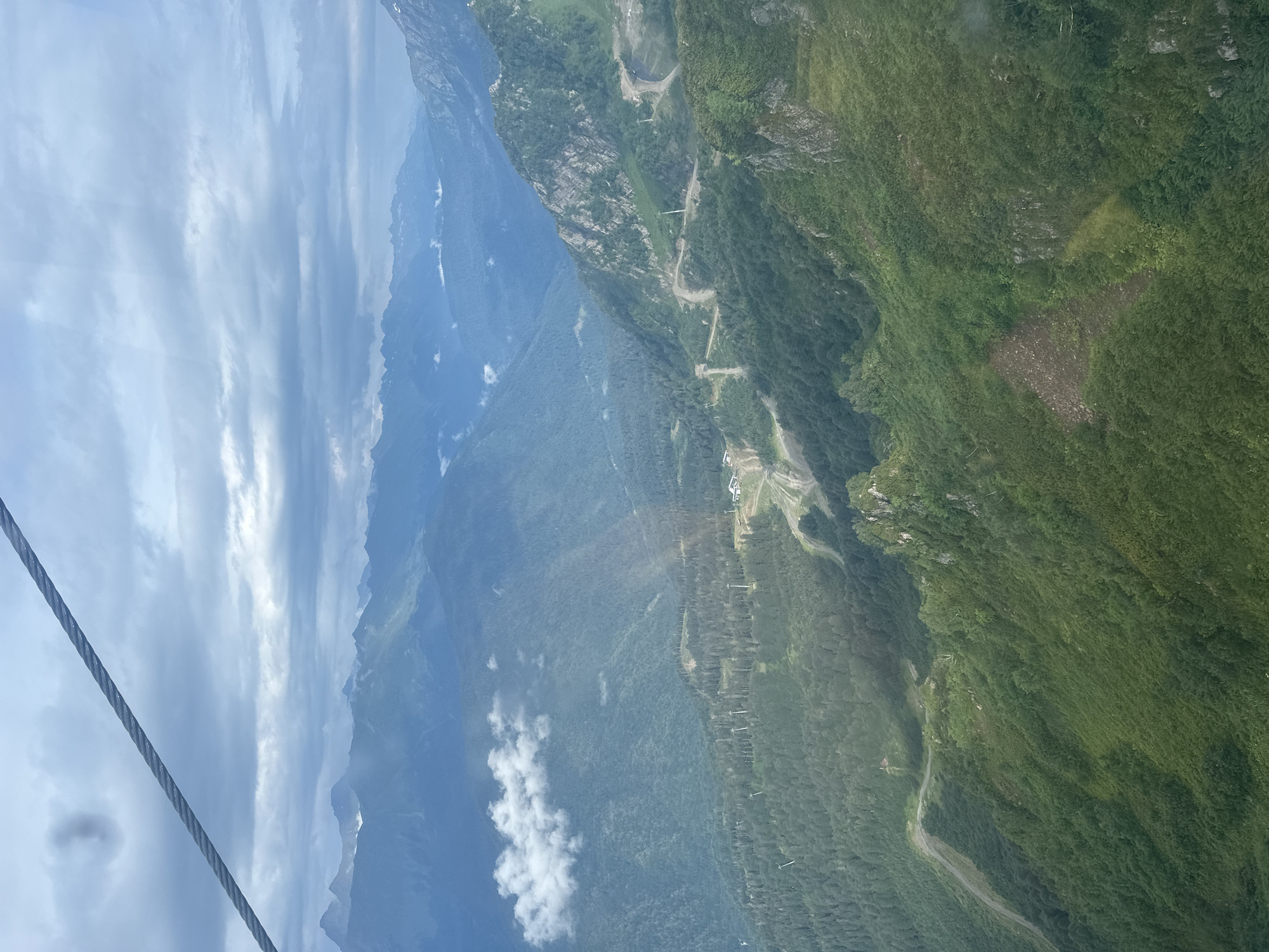 The image captures a panoramic view of a mountainous landscape, likely taken from a cable car. In the foreground, a thick steel cable stretches diagonally across the upper left quadrant of the frame, suggesting the vantage point. The scene is dominated by rolling green mountains covered in dense forests. Several winding dirt roads or trails are visible, snaking their way up the mountainsides. Scattered throughout the scene are numerous ski lift pylons, indicating this is a ski resort area. In the distance, layers of further mountain ranges recede into a hazy, overcast sky. A few wispy clouds drift amongst the peaks. The overall impression is one of a vast, natural, and somewhat wild mountainous region, likely an active outdoor recreational area. The location is identified as Estosadok, Russia, a known ski resort town in the Krasnaya Polyana region.