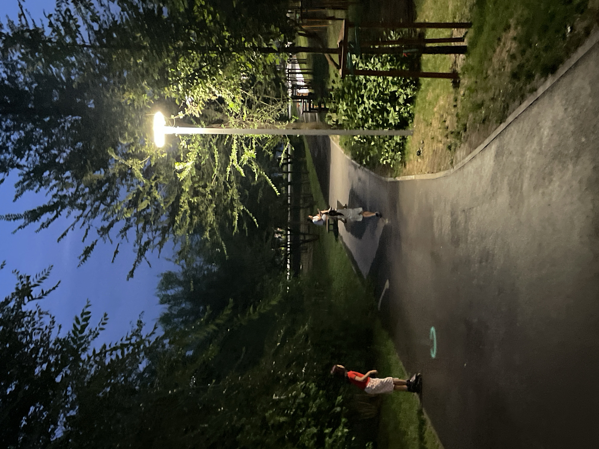 The image depicts a park-like setting at dusk or nighttime, illuminated by a lamppost. A paved pathway curves through the scene, bordered by grass and trees. In the foreground on the left, a young person wearing a red t-shirt, light-colored shorts, and rollerblades is standing on the pathway. A glowing green circle is visible on the ground near their feet. Further along the pathway, two other figures are present. One, appearing to be a child in a white outfit, is walking away from the camera. Another figure, partially visible, is seated on a bench. The lamppost is a prominent feature, casting light on the surrounding vegetation and pathway. The sky is a deep blue, indicating the time of day. The location is identified as Almaty, Kazakhstan.