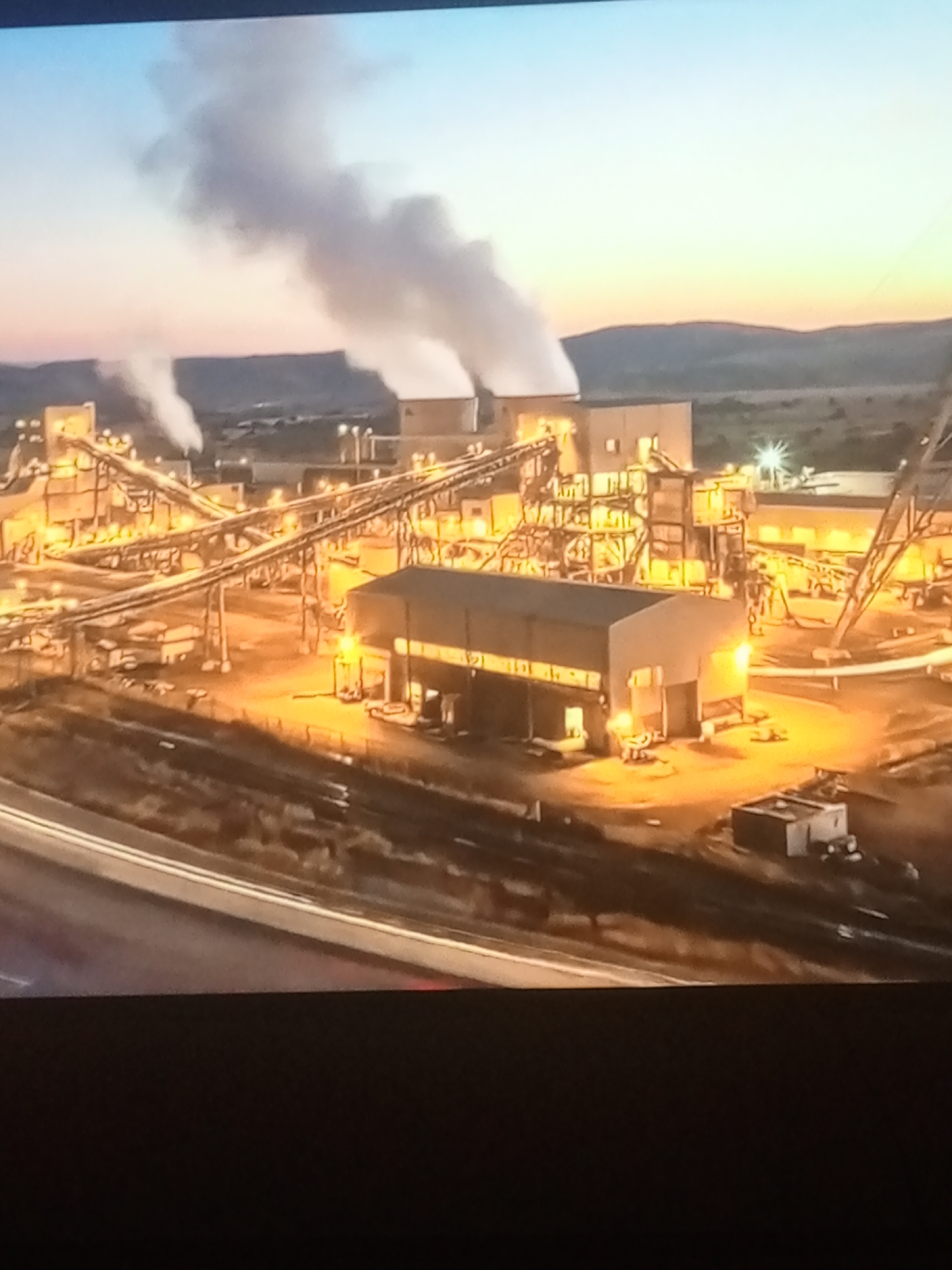 An industrial complex is pictured at dusk, emitting plumes of white smoke into the sky. The scene is lit by the warm glow of artificial lights illuminating the structures and conveyor belts. In the foreground, a highway runs horizontally across the frame, with a dark, out-of-focus area at the bottom of the image. The background consists of rolling hills under a twilight sky transitioning from a soft orange near the horizon to a light blue above. The industrial site features multiple buildings, large metal structures, and what appear to be processing or mining machinery. There are no visible people or vehicles in the image.