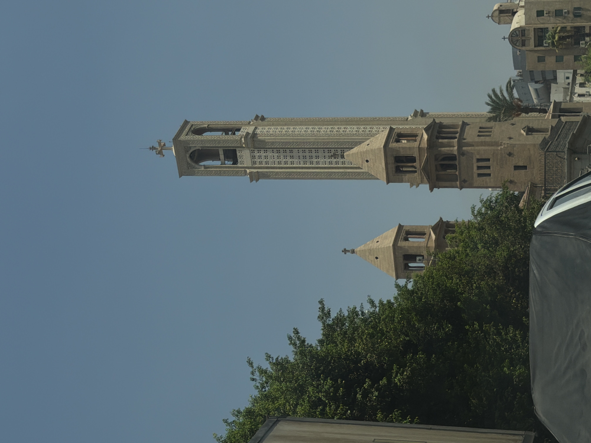 The image captures a view of the Saint Mark's Coptic Orthodox Cathedral in Cairo, Egypt. Two prominent bell towers are visible, one taller and more ornate than the other. The taller tower is square-based and features intricate decorative patterns along its height, with arched openings near the top where bells would be located. It is topped with a cross. The shorter tower is octagonal at its base and has a conical roof, also topped with a cross.
In the foreground, a portion of a vehicle's roof is visible, covered with a dark tarp. To the left of the towers, a dense, leafy green tree obscures some of the lower parts of the scene. Behind the shorter tower and to the right, other buildings of varying architectural styles can be seen, including a structure with a distinctive arched facade and crosses, hinting at the presence of other religious or historical buildings within the vicinity. A palm tree is also visible to the right of the shorter tower. The sky is a clear, pale blue, suggesting a sunny day. The overall scene depicts an urban environment with a strong religious architectural presence.