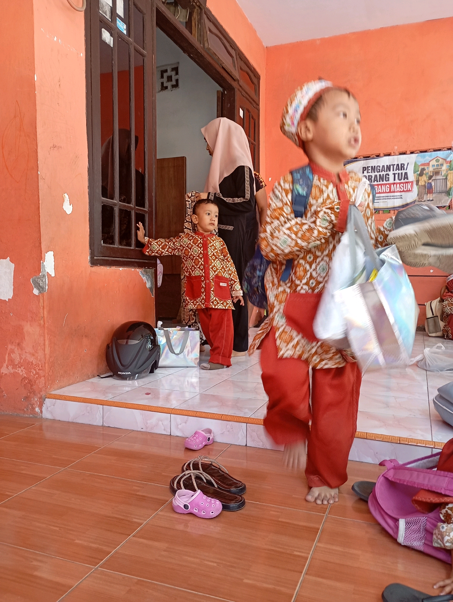 A young boy in a patterned shirt and red pants, wearing a matching hat, stands on a tiled porch in front of an orange building. He is looking to the right and holding a shiny silver bag. Another young boy in a similar outfit stands near a window to the left, with his hand on the window frame. A woman in a light pink hijab and black patterned dress stands behind the first boy, and another woman in a dark hijab is visible in the reflection of the window. A black helmet is on the porch floor, along with a white and silver tote bag. Several pairs of shoes are scattered on the floor, including pink children's sandals and brown sandals. A sign with text and images is visible on the wall in the background. The environment appears to be an outdoor area in Ketegan, Indonesia.