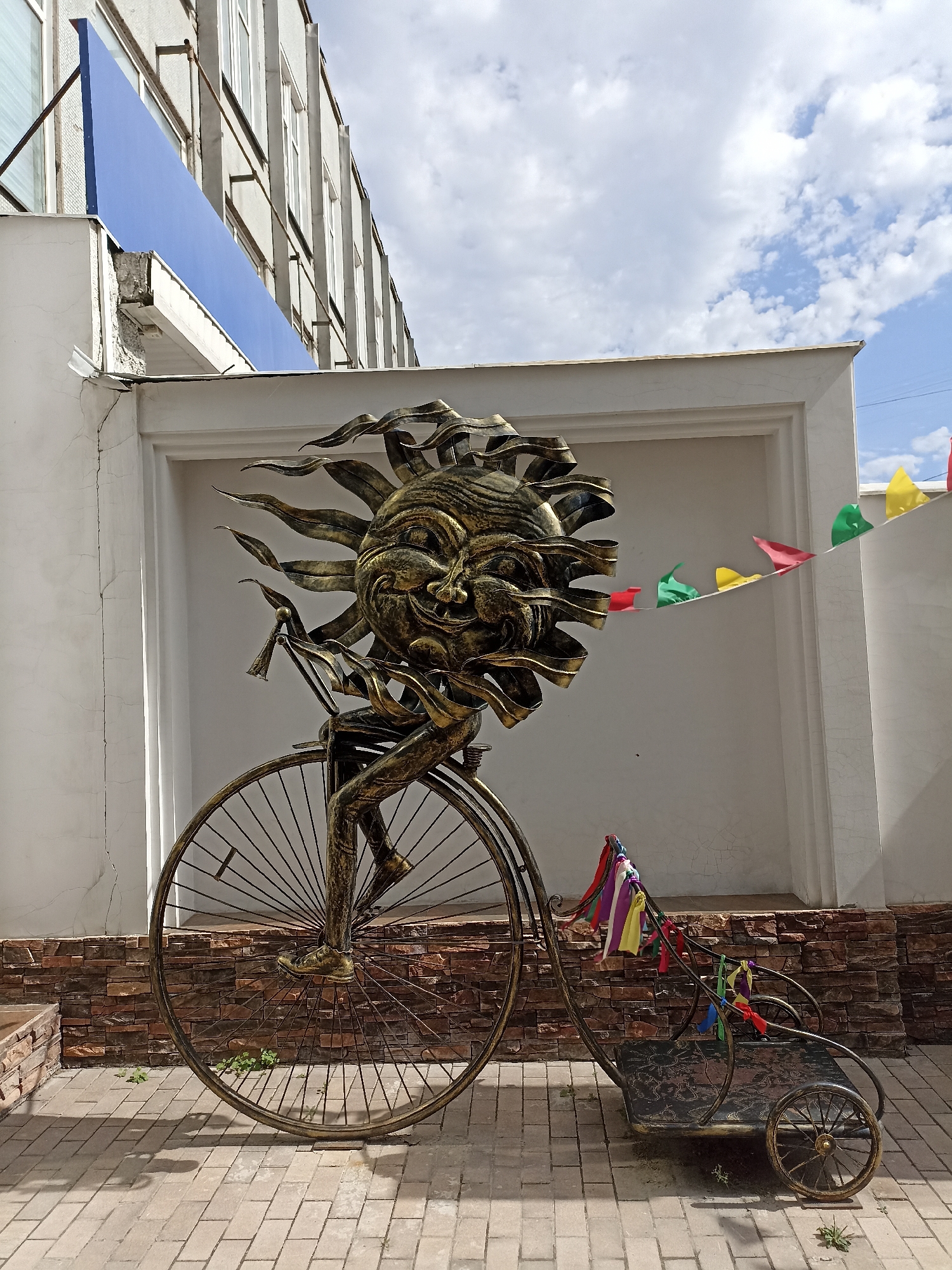 This is a full, eye-level, outdoor medium shot of a metallic sculpture of a sun with a face, riding a bicycle, located in front of a building on a sunny day. The sculpture is gold and bronze in color, and it is intricately detailed, with flames coming out of the sun's head. The sun's face is smiling and has a mischievous expression. The sun is riding a bicycle with a large front wheel and a smaller back wheel. There is a small cart attached to the back of the bicycle. The cart is decorated with colorful ribbons. In the background, there is a building with white walls and a blue sign. There are also a string of colorful flags hanging on the wall. The ground is paved with bricks. The sculpture is located in Novosibirsk, Russia.
