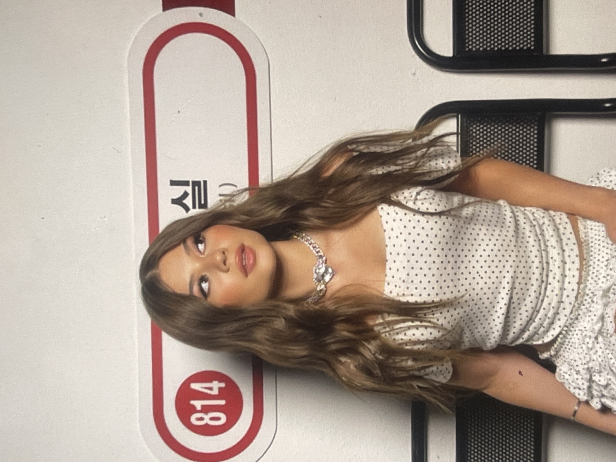 A young woman with long, wavy brown hair, fair skin, and full makeup is captured in an indoor setting. She is wearing a white top with black polka dots and a matching skirt or shorts. A notable silver-colored chain necklace with a large, ornate, possibly heart-shaped jeweled pendant is visible around her neck. She poses looking upwards and to her left.
To her left, a prominent vertical white sign with a thick red border features a red circle at the bottom containing the numbers "814". Above this, vertically oriented Korean characters are visible, specifically "실" and a partial character that appears to be "ㅂ" or "ㅃ". To her right, a black metal structure with a grid pattern, possibly part of a bed frame or shelving, is present. The image itself is rotated 90 degrees clockwise. The scene is brightly lit, suggesting daytime or a well-lit interior. The provided location context is Poltava, Ukraine, despite the presence of Korean text on the sign.