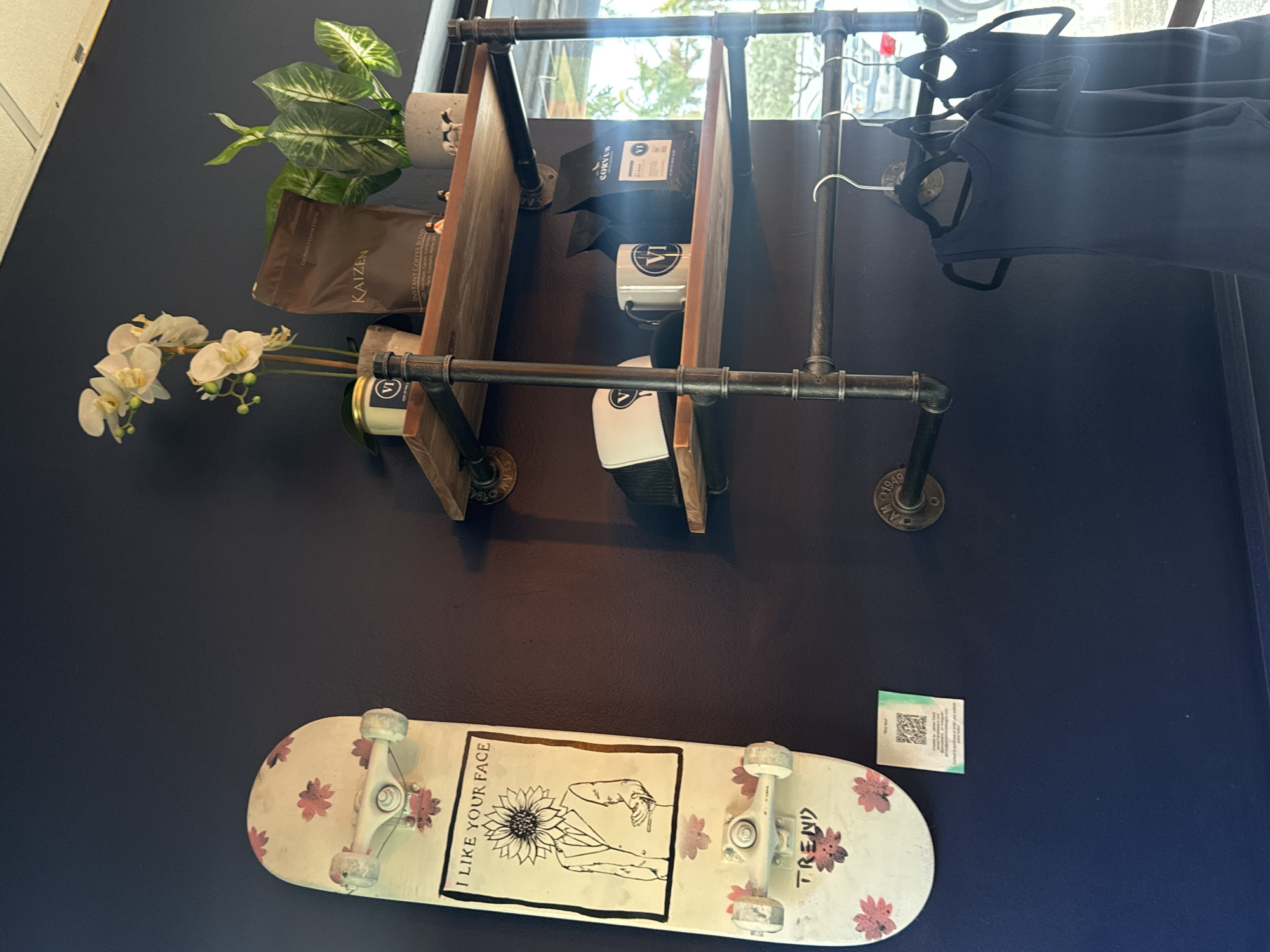 Here is a description of the image:
A dark blue wall in Boca Raton, Florida features a metal and wood shelf system. The shelves hold a plant, a bag of Kaizen Instant Coffee Blend, a small candle, Corvus coffee bag, a white coffee mug with a black logo (LA), and a white bottle with a black logo (LA). A skateboard, adorned with a sunflower and the words "I LIKE YOUR FACE" and "TREND", is mounted on the wall below the shelf. A small, rectangular QR code sticker sits to the right of the skateboard. Clothing hangs to the right of the shelves on hangers. White orchids are placed at the left of the shelves. The shelves are constructed with iron pipe.