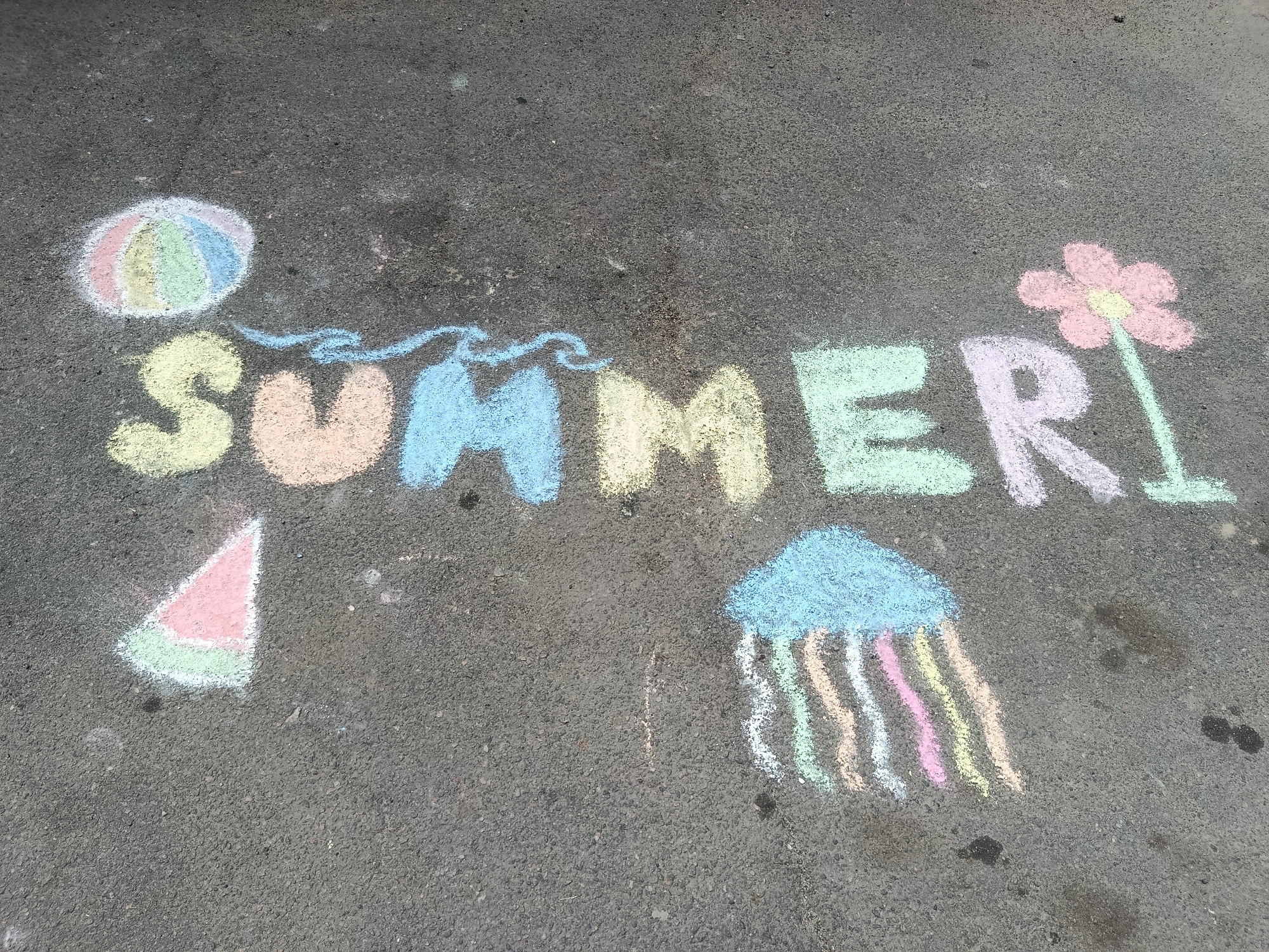 Chalk drawings on asphalt depict the word "SUMMER" in colorful, blocky letters. To the left of the word, a beach ball is drawn in pastel shades of pink, yellow, green, and blue. A wavy blue line connects the ball to the first letter "S." Below the word, a slice of watermelon is depicted in green, white, and pink. To the right of the word, a pink flower with a yellow center and a green stem is drawn. Below the flower and to the right of the word, a light blue jellyfish with colorful tentacles in white, peach, green, and pink is illustrated. The drawings are on a dark gray asphalt surface with some visible smudges and debris. The scene appears to be outdoors in an urban environment.
