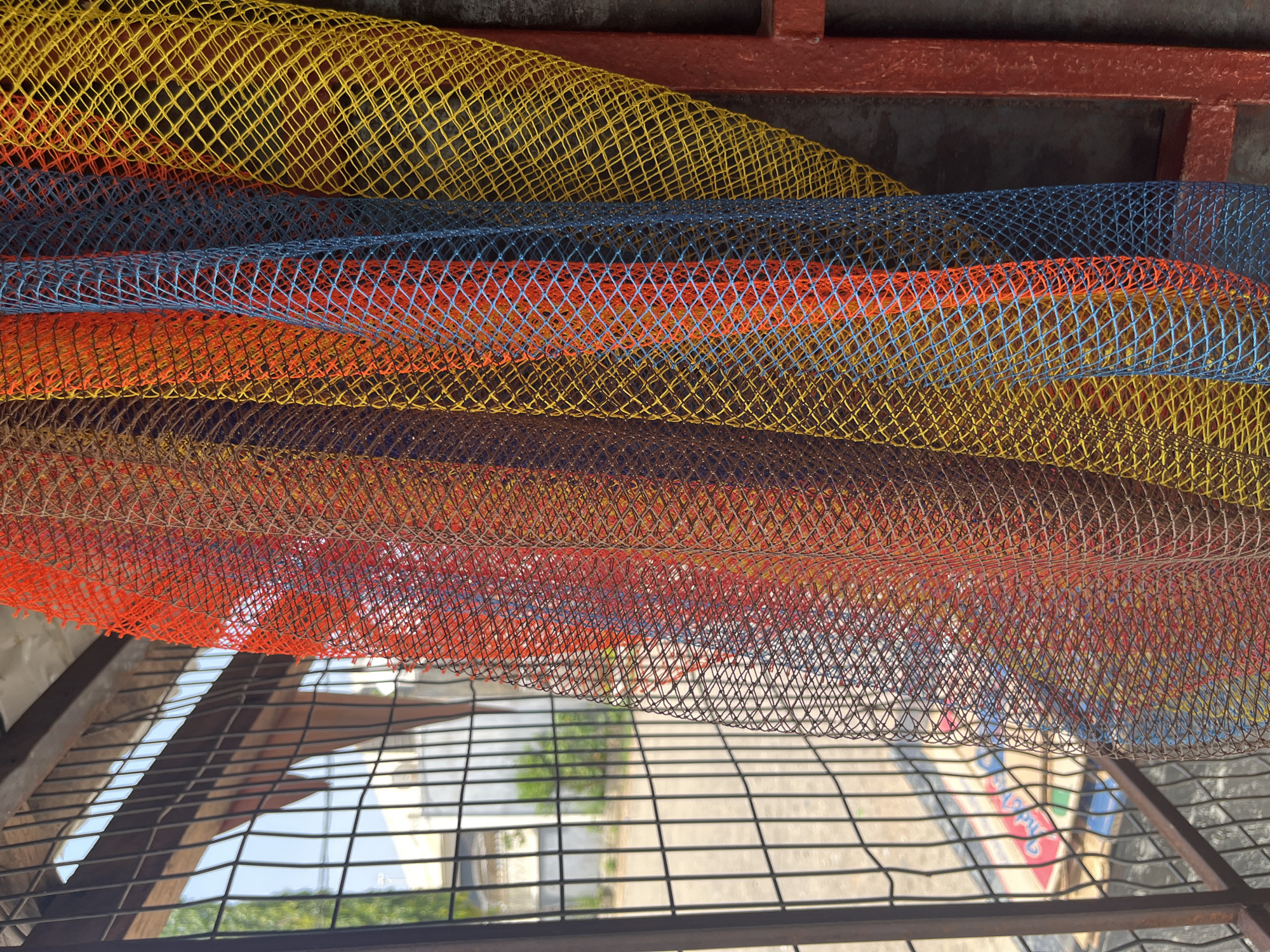 This is a close-up, eye-level shot of a fence on a sunny day, likely in Abuja, Nigeria. The fence is made of dark metal bars and a wire mesh. Hanging in front of the fence are several colorful netting materials. The netting is draped and layered, creating a visual texture. The colors visible in the netting are red, orange, blue, and yellow. The background shows a glimpse of a building and trees through the fence, suggesting an outdoor urban environment. The lighting is bright and appears to be natural daylight.