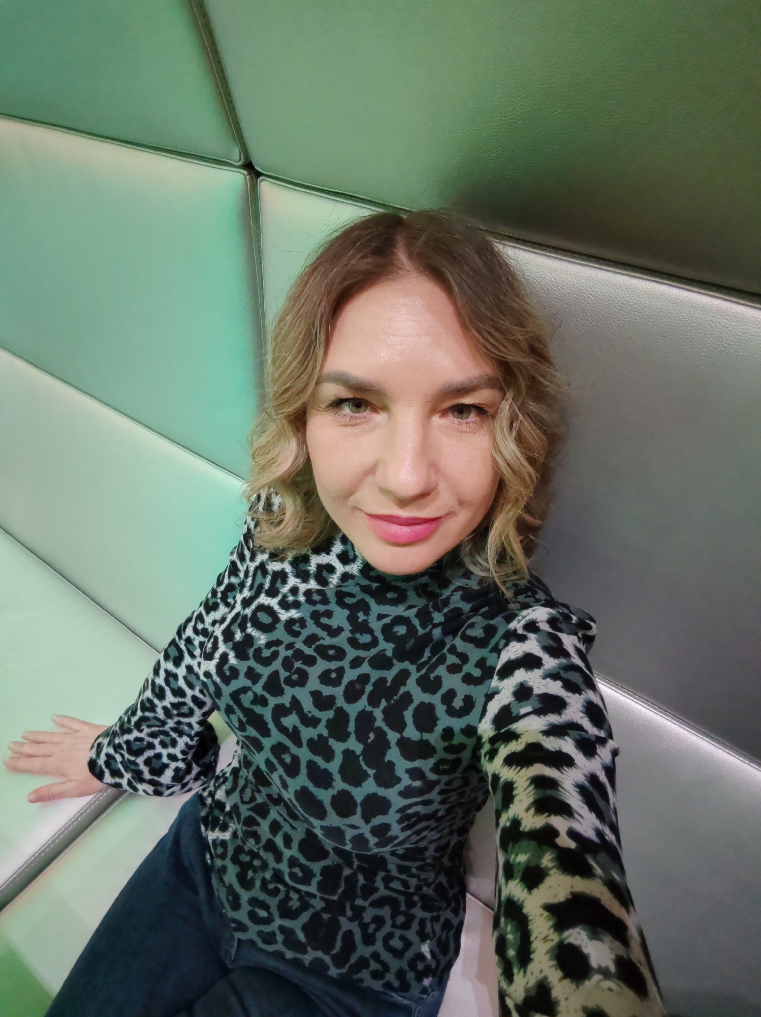A woman poses for a selfie indoors, sitting on a modern, padded bench or seating area. She is positioned against a geometrically paneled wall or backrest, which has a glossy or leather-like texture and reflects a green-blue hue, possibly from ambient lighting.
The woman has blonde, wavy hair and is looking directly at the camera with a slight smile. She is wearing light makeup, including pink lipstick, which contrasts with her green or hazel eyes. Her attire consists of a long-sleeved turtleneck top with a teal/green and black leopard print pattern, paired with dark blue jeans. One of her hands is visible, resting on the padded seat beside her. The overall impression is a casual, contemporary portrait, likely taken in a lounge, bar, or a similar indoor public space. There is no visible text in the image.