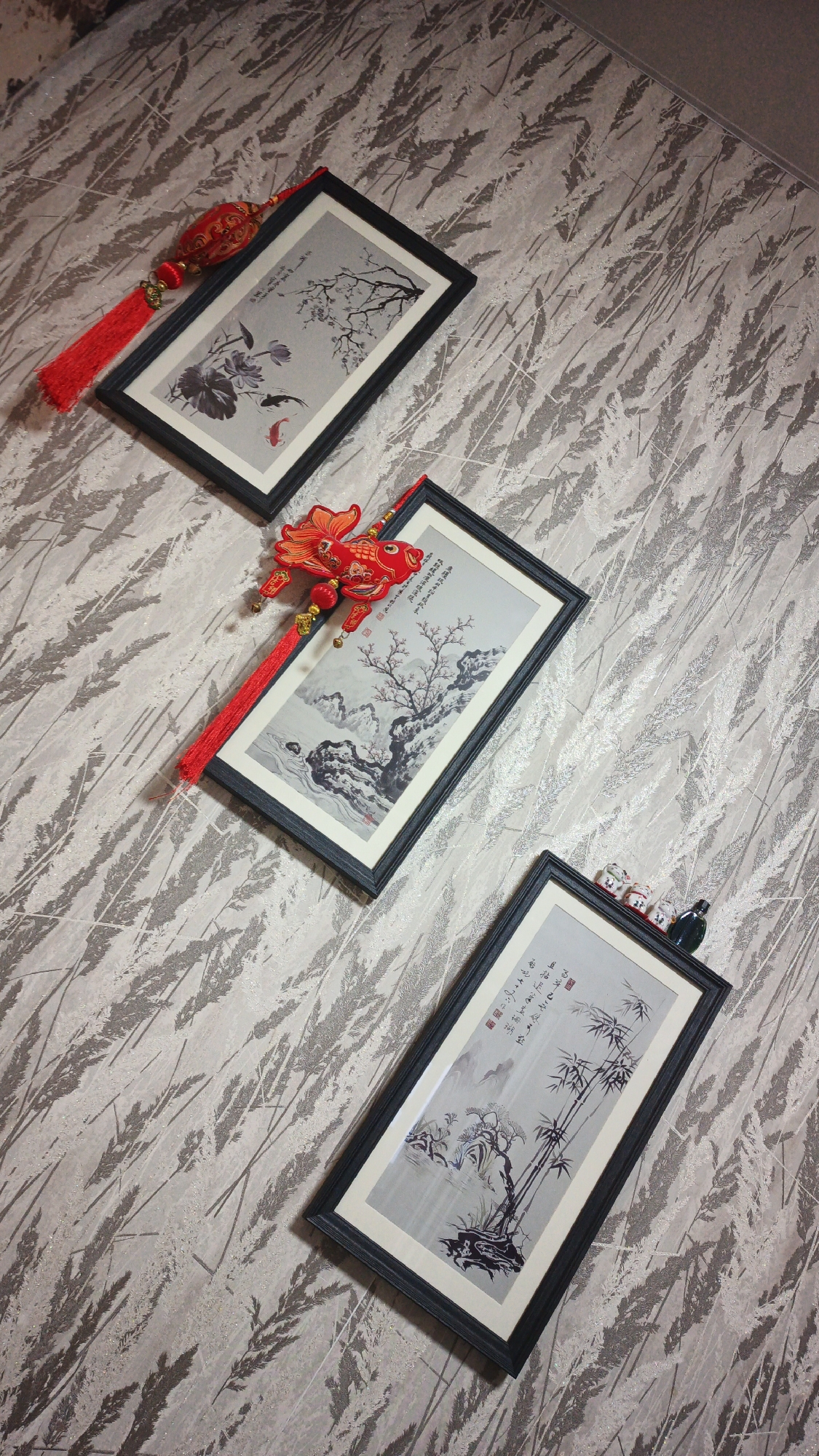 Three framed Chinese paintings are displayed diagonally on a textured wall. Each painting features traditional ink wash artwork and is adorned with red Chinese New Year ornaments. The top painting depicts lotuses and koi fish, with a red lantern and tassel hanging from its frame. The middle painting shows a mountainous landscape with a bare tree, also decorated with a red fish ornament and tassel. The bottom painting displays bamboo and rocks, with small decorative figures and a dark green accent on its frame. The wall has a repeating pattern of stylized leaves or reeds in shades of gray and white. The scene is brightly lit, and no people are present. The location is the city of Drezna, Russia.